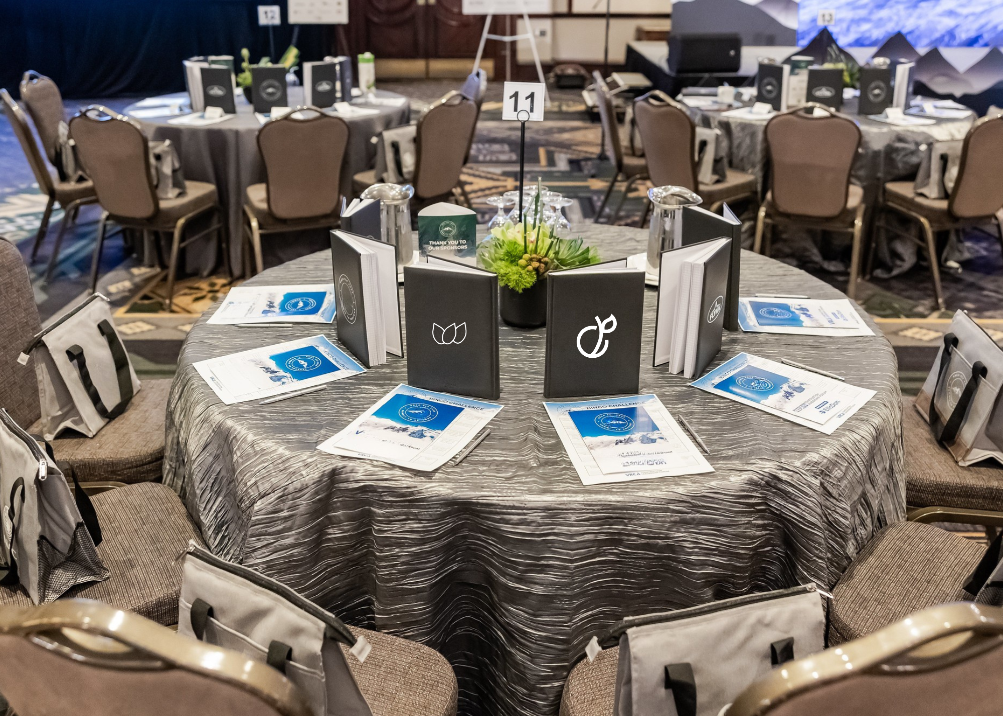 Round banquet table with blueprints, menu cards, and a floral centerpiece at the center, set for a formal event at a conference hall.