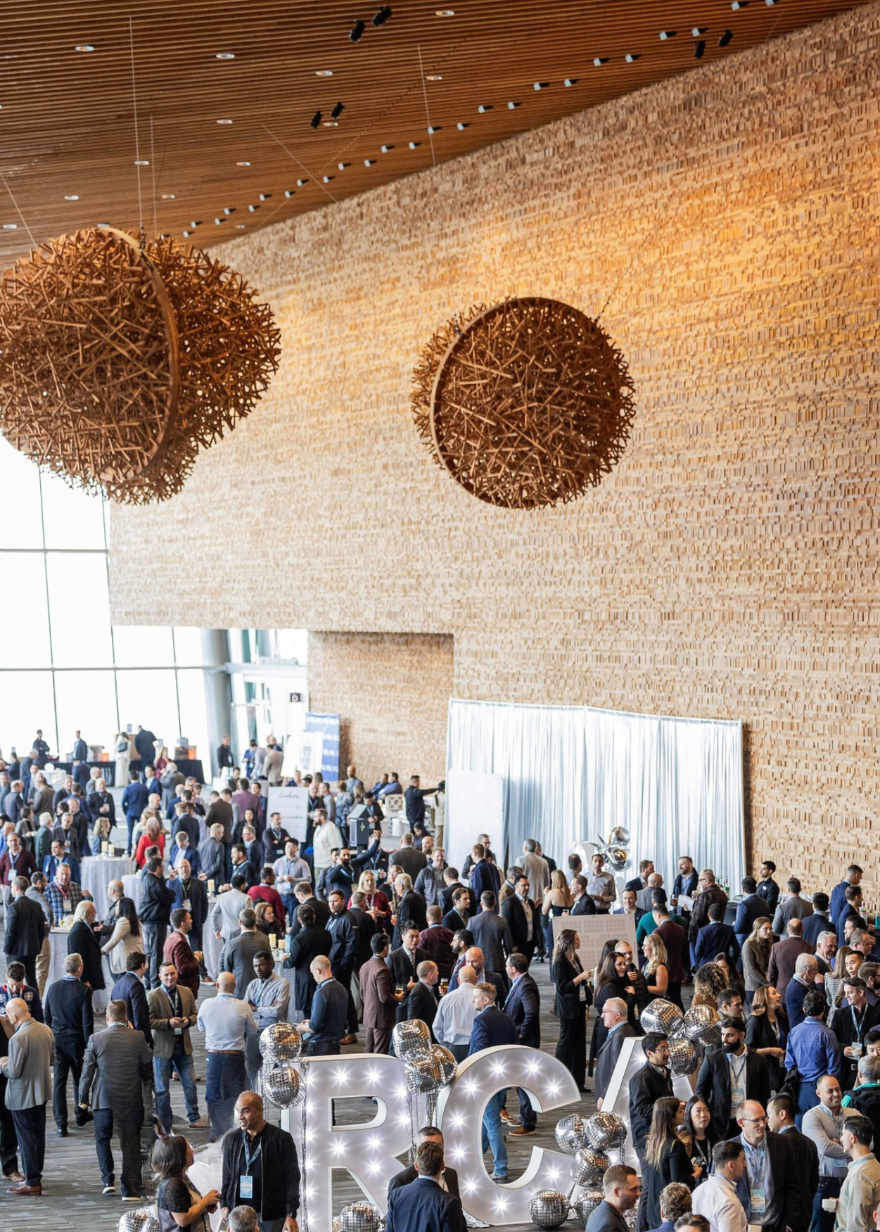 Large conference or networking event with many attendees, crowded room, people engaging in conversations and activities, front displays with illuminated letters and balloons, wooden wall decor, large window letting in natural light.