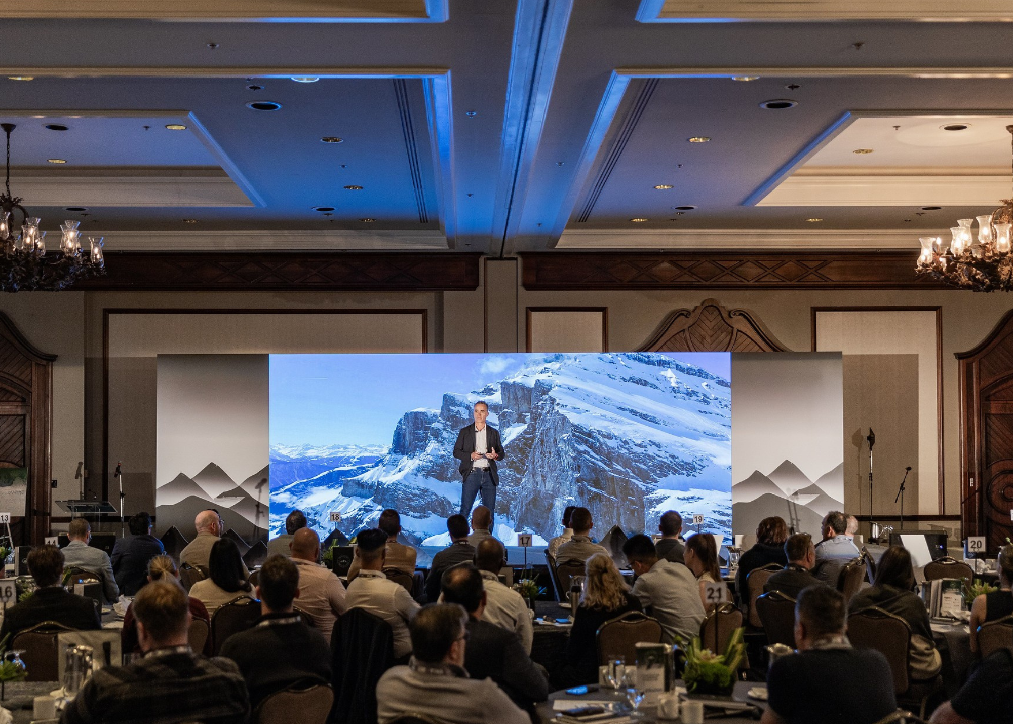 A speaker presenting at a conference with a large mountain background on the screen behind him, audience seated at tables in a formal conference room.