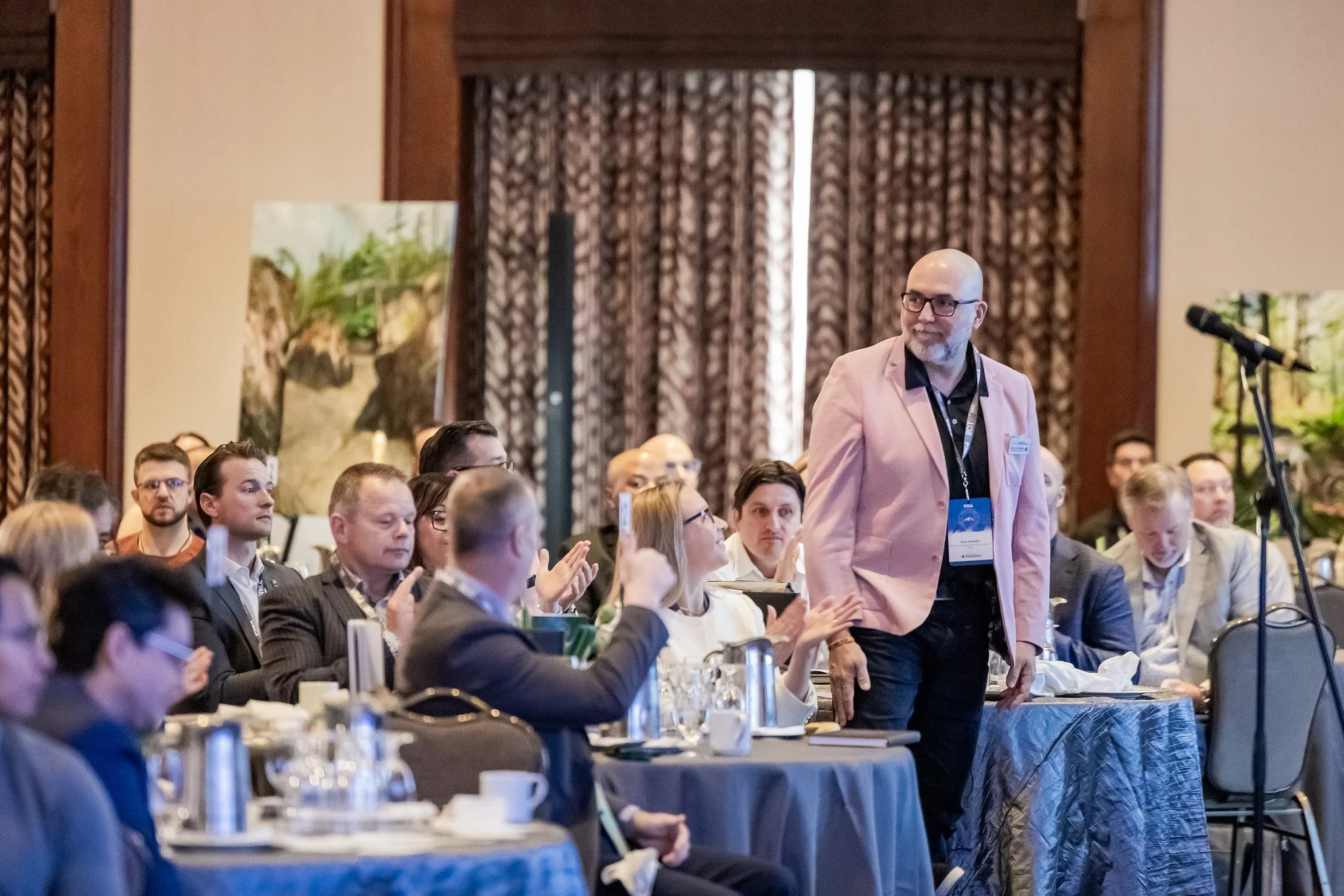 A man in a pink blazer walking past a seated audience during a conference or seminar in a large room with patterned curtains and artwork.