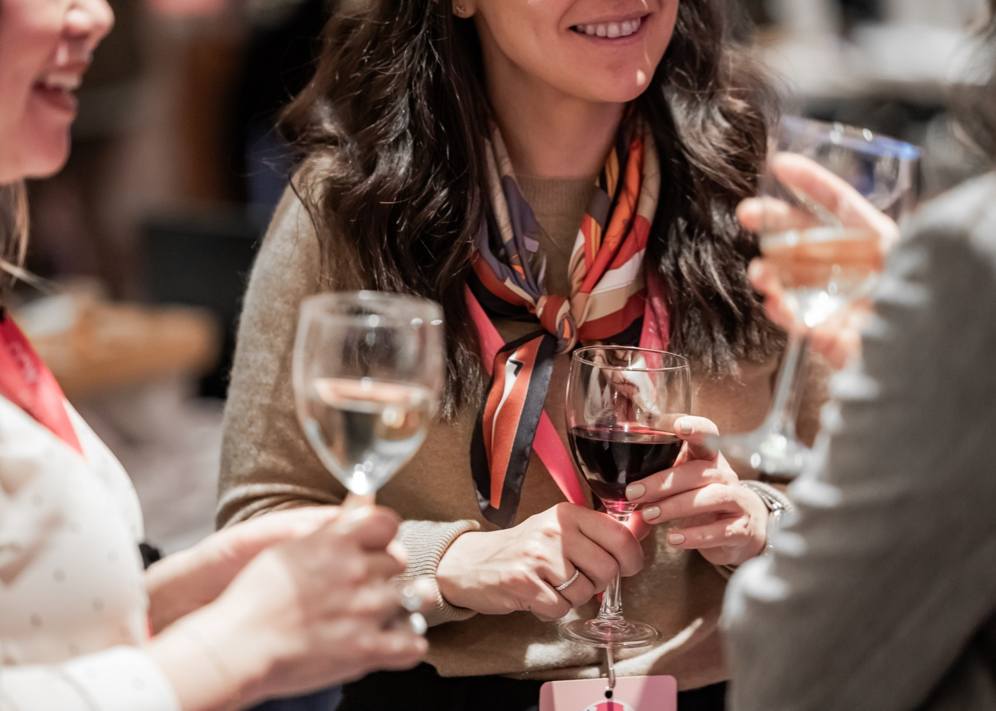 Three women enjoying drinks at a social gathering, with one woman holding a glass of red wine and others holding glasses of water or cocktails, engaging in conversation.