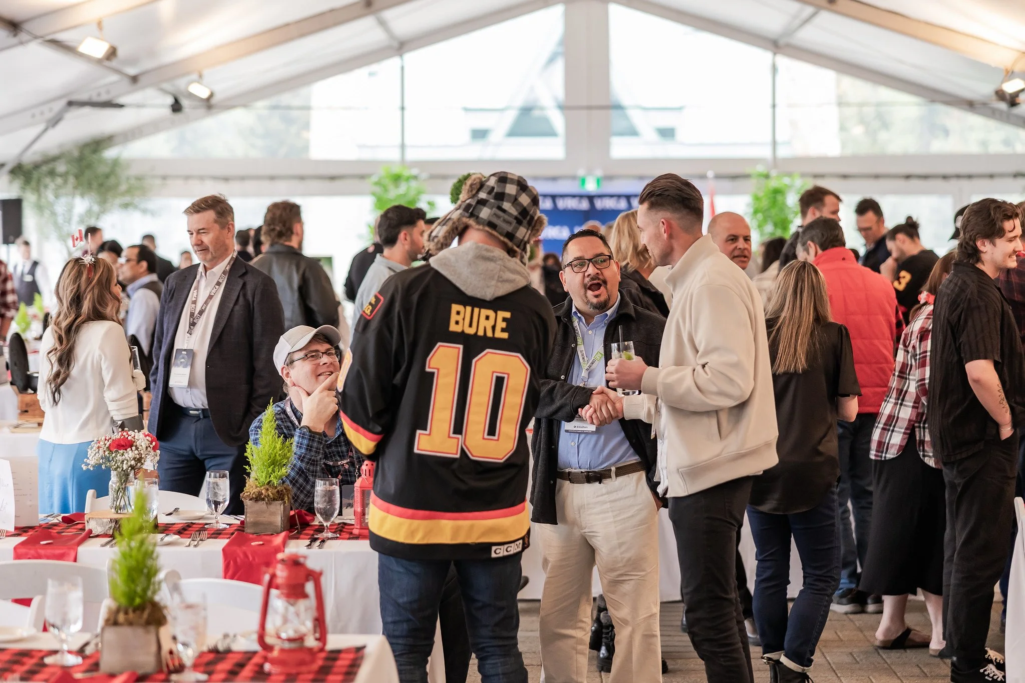 Group of people socializing at a gathering, with some seated at tables and others standing and talking, inside a large tent with natural light.