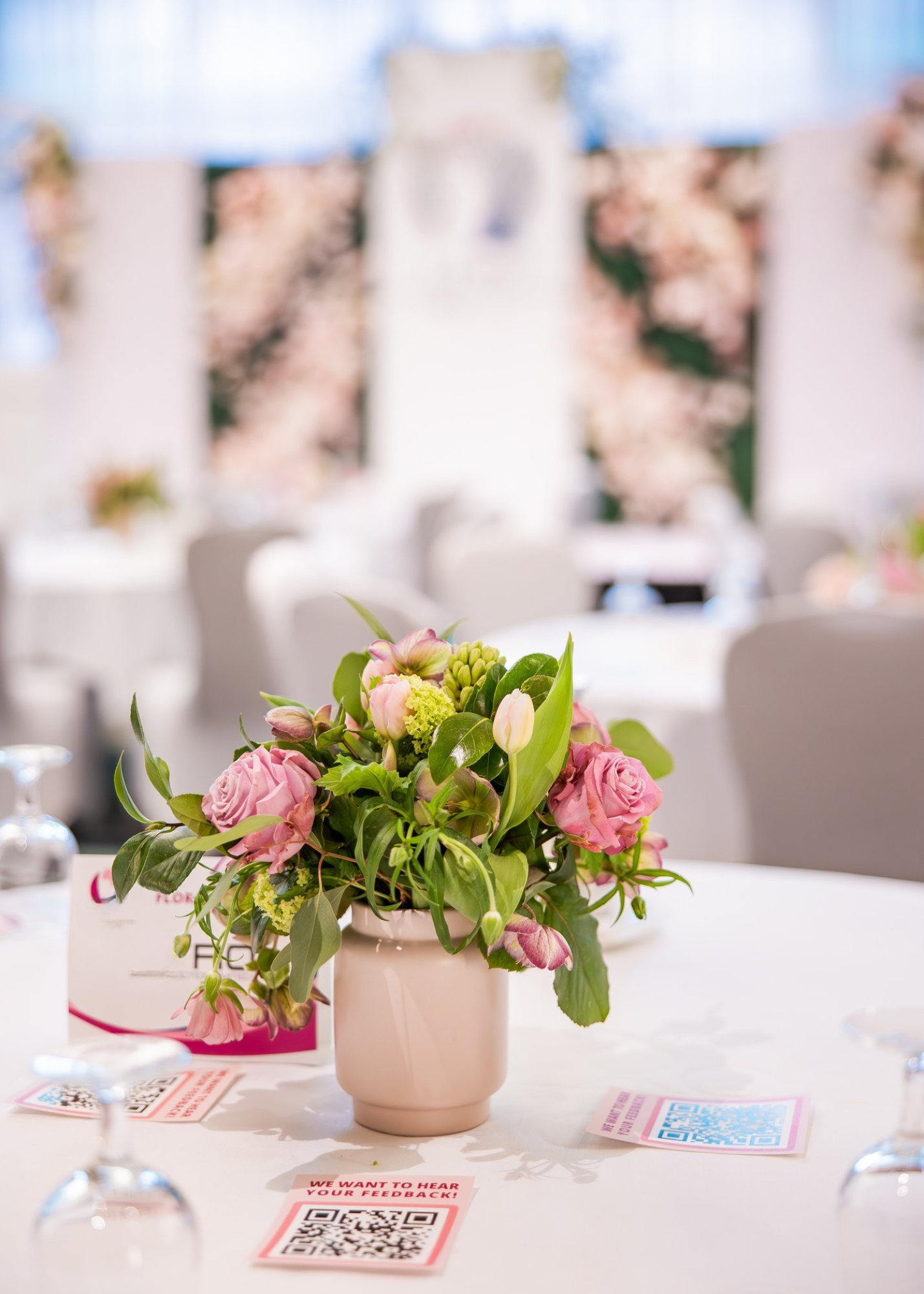 A pink floral centerpiece with roses, tulips, and greenery on a white table at a wedding or event reception.