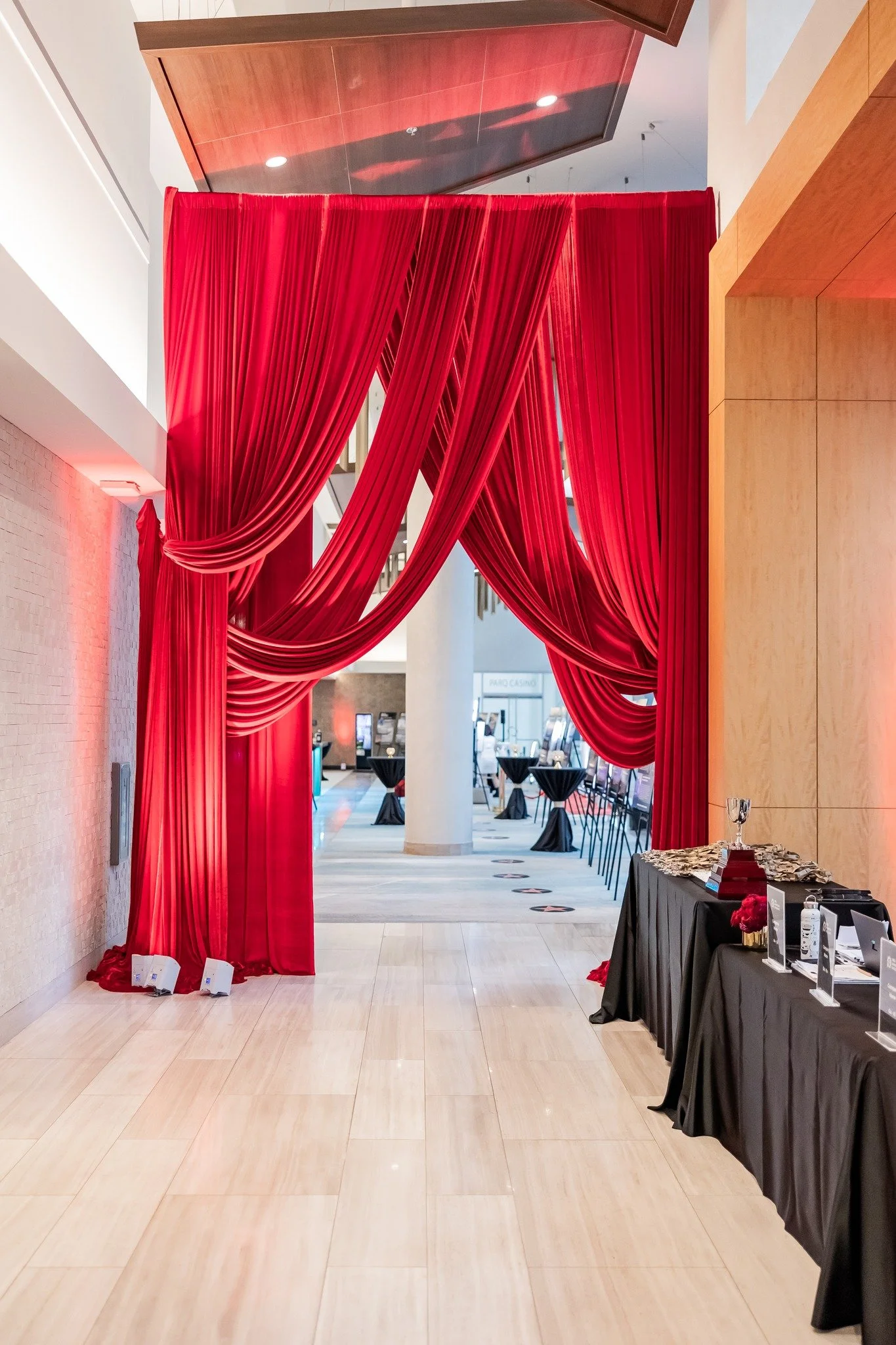 An elegant indoor installation with red curtains creating a grand entrance, black tablecloth-covered tables on the right adorned with a wine glass and decorative items, in a modern venue.