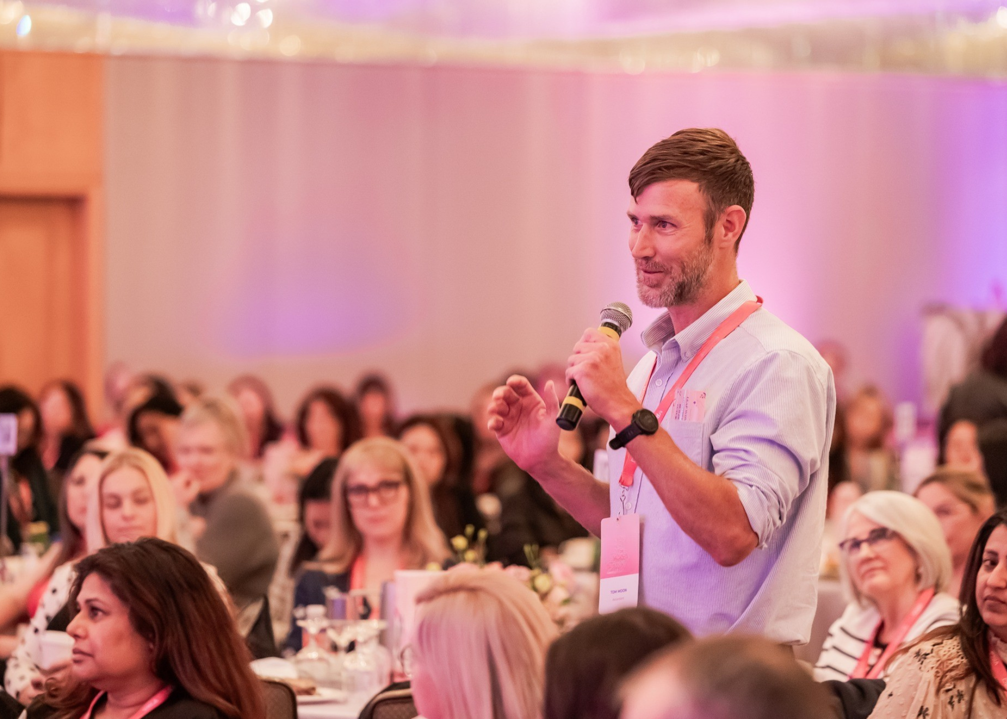 A man with brown hair and a beard holding a microphone in a conference room filled with women seated at tables.