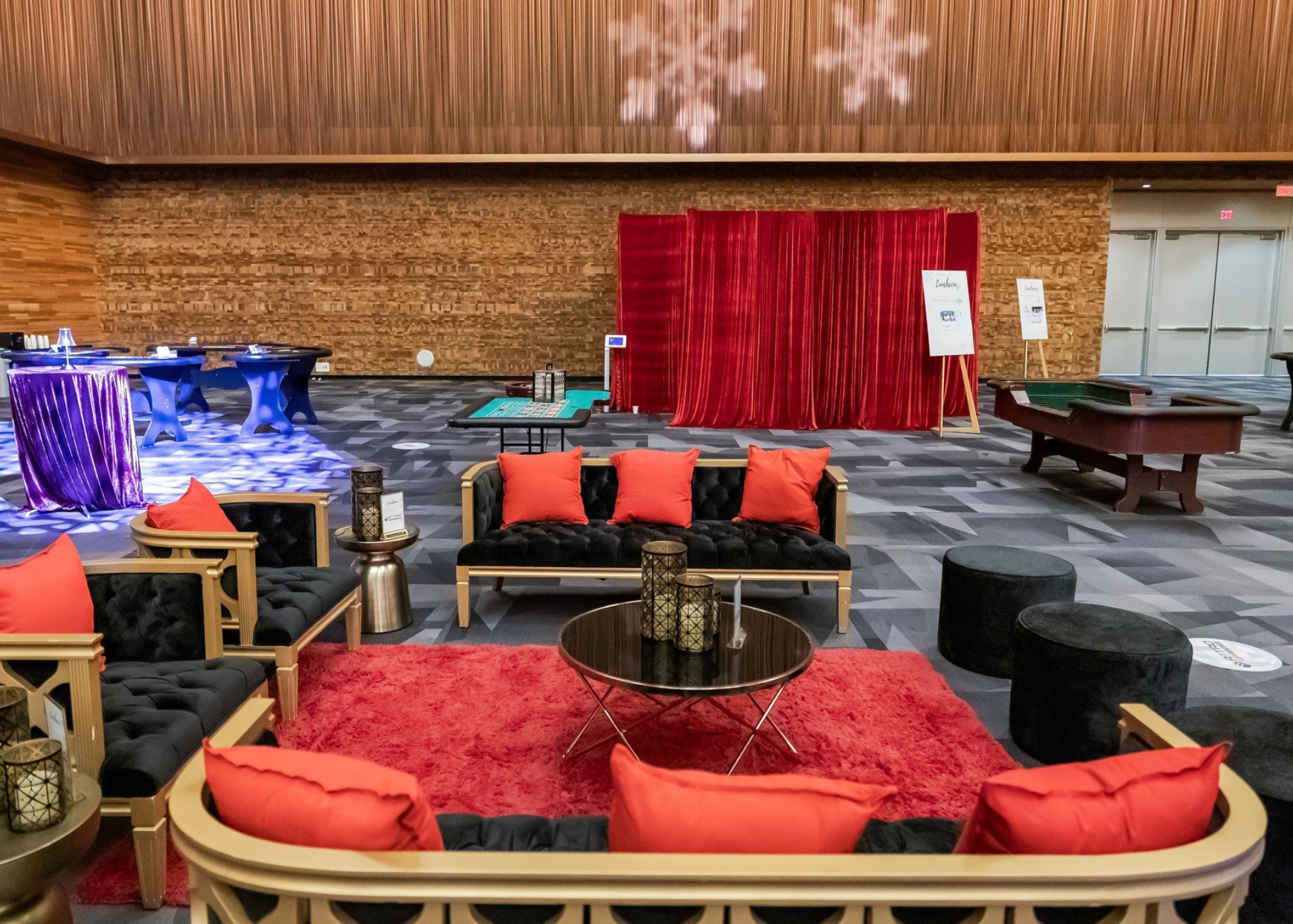 A hotel lobby lounge with black and gold furniture, red pillows, a red rug, and black ottomans. In the background, a red velvet curtain and a bar area with blue-lit tables and purple tablecloths are visible.