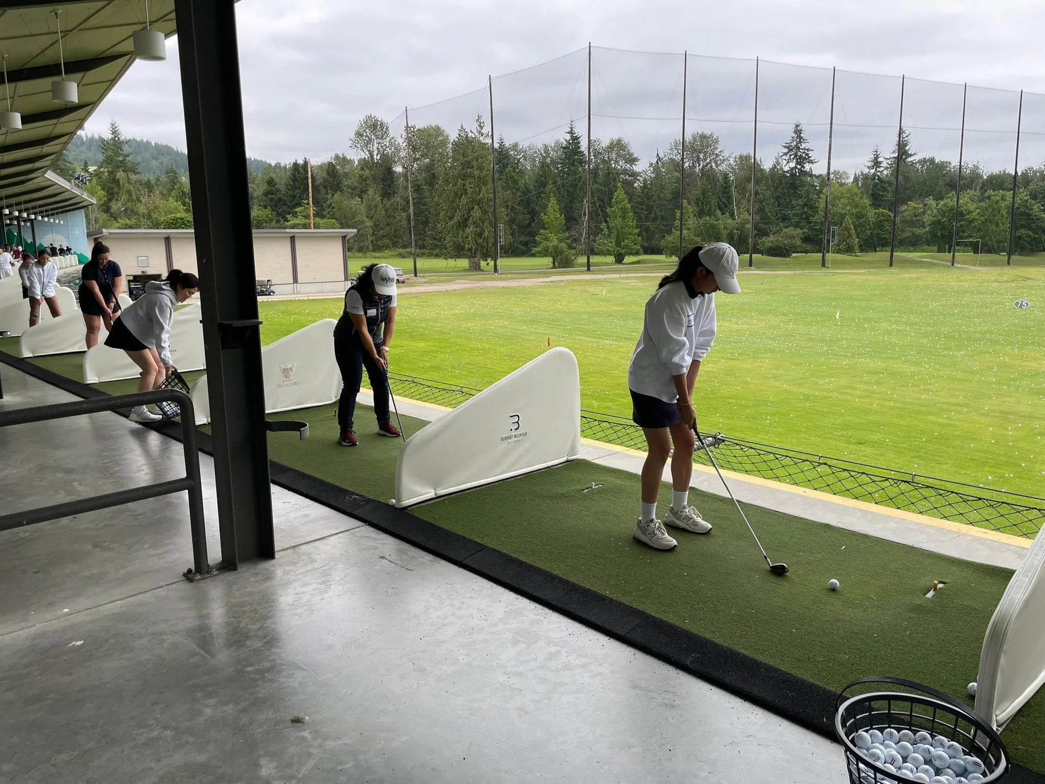 Group of people practicing golf at an indoor driving range on a rainy day, with a view of a green golf course outside.