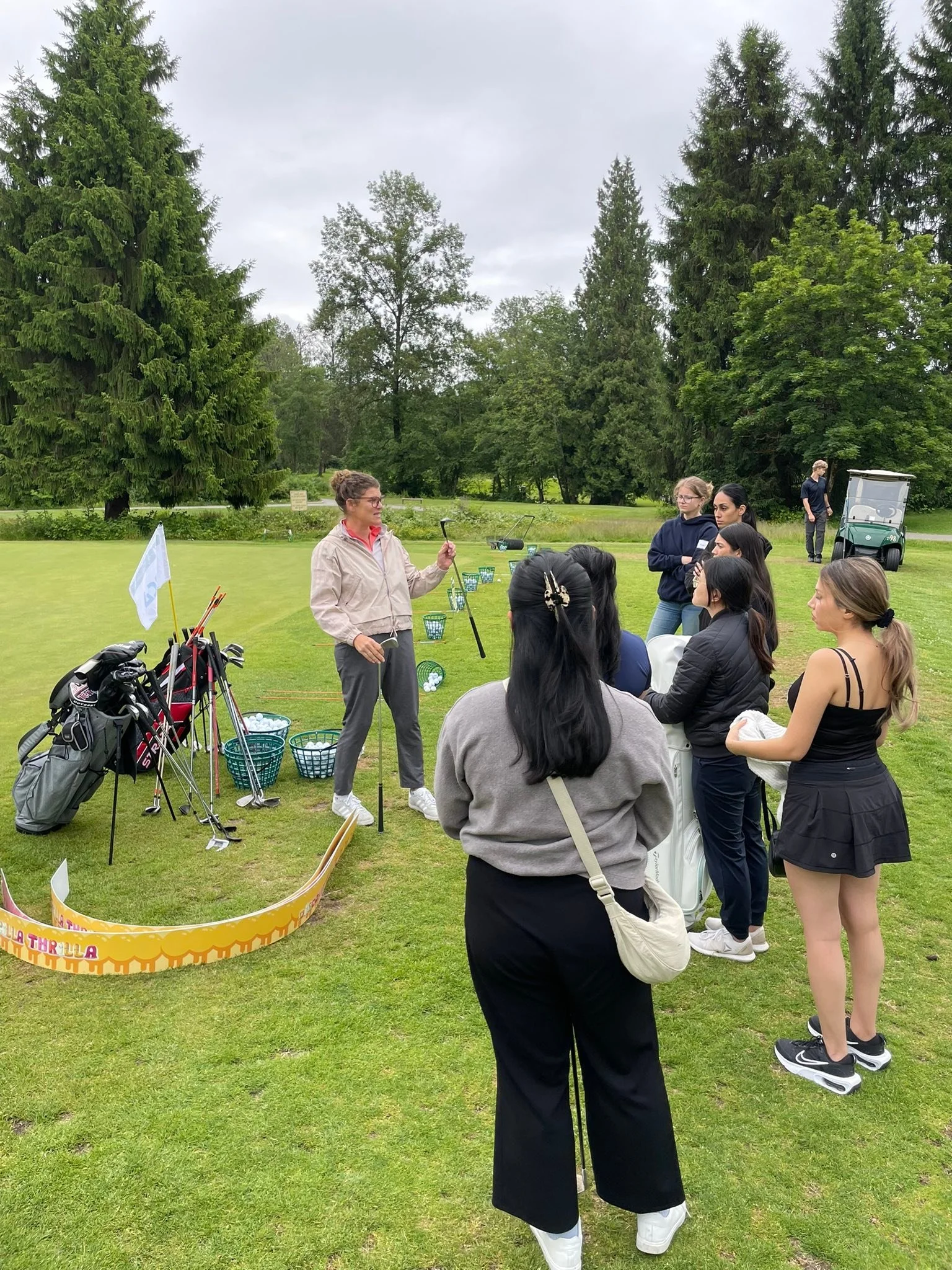 A group of women and girls listening to a golf instructor on a golf course surrounded by trees, with golf bags, clubs, and baskets nearby, and a golf cart in the background.