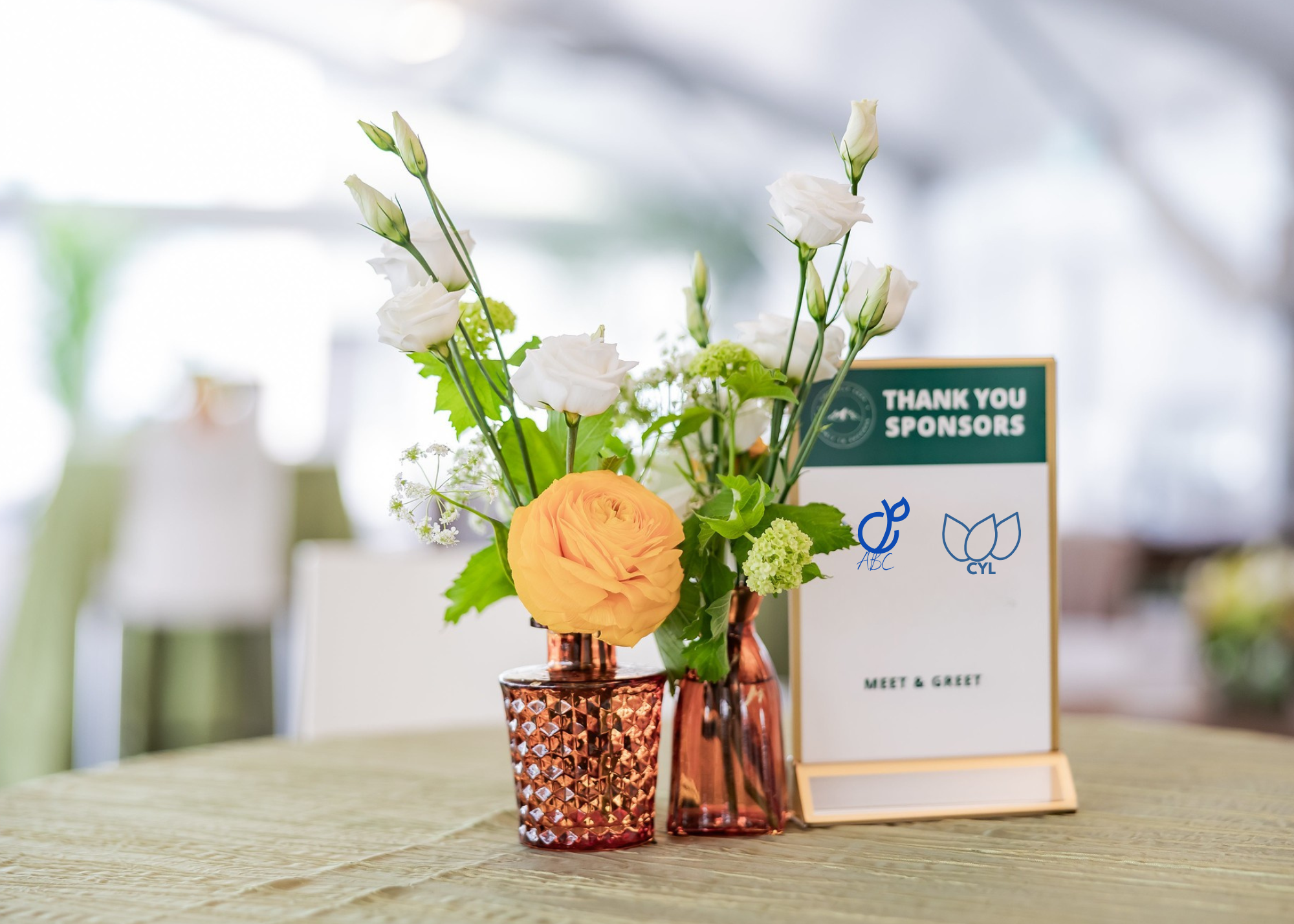 A floral arrangement with white and peach flowers in pink glass vases on a table, alongside a sign thanking sponsors for a meet and greet event.