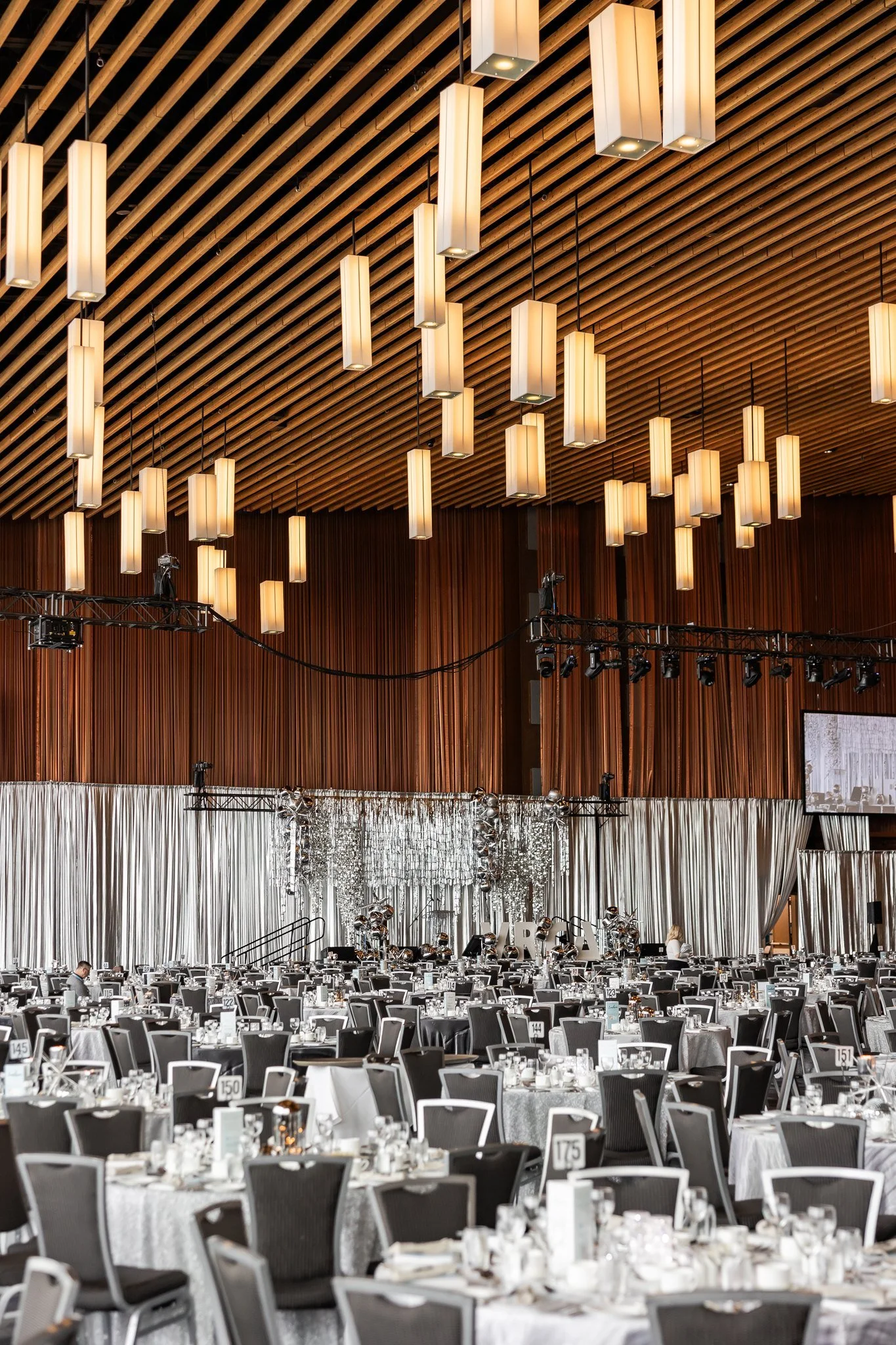 A large banquet hall decorated with multiple round tables set for an event, with silver and white drapery backdrop and hanging lights from a wooden ceiling.
