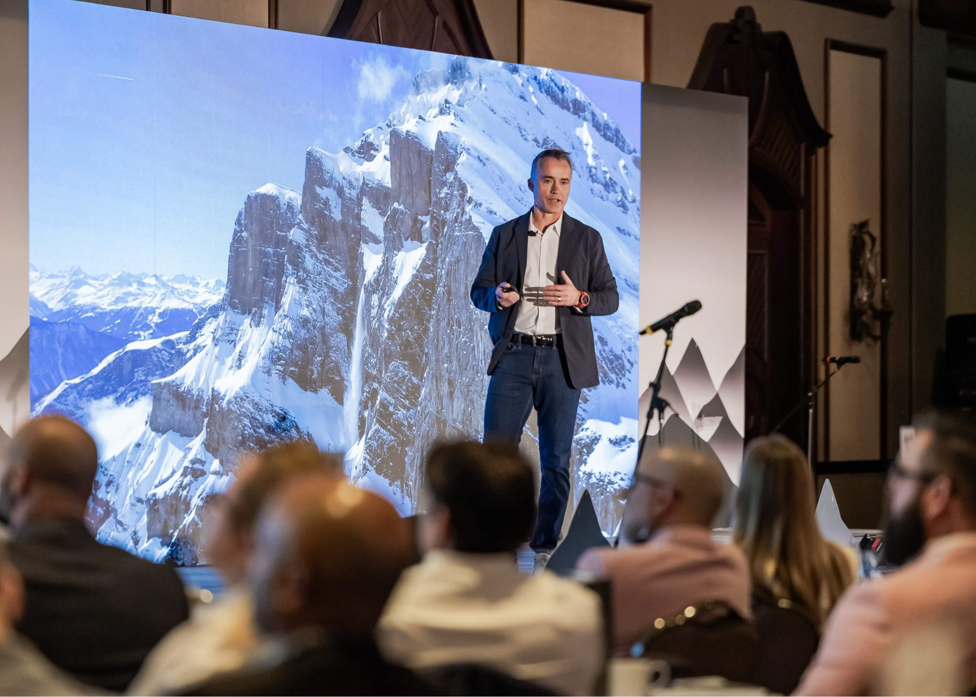 A man giving a presentation on stage with a large mountain landscape background behind him, audience members sitting and listening.