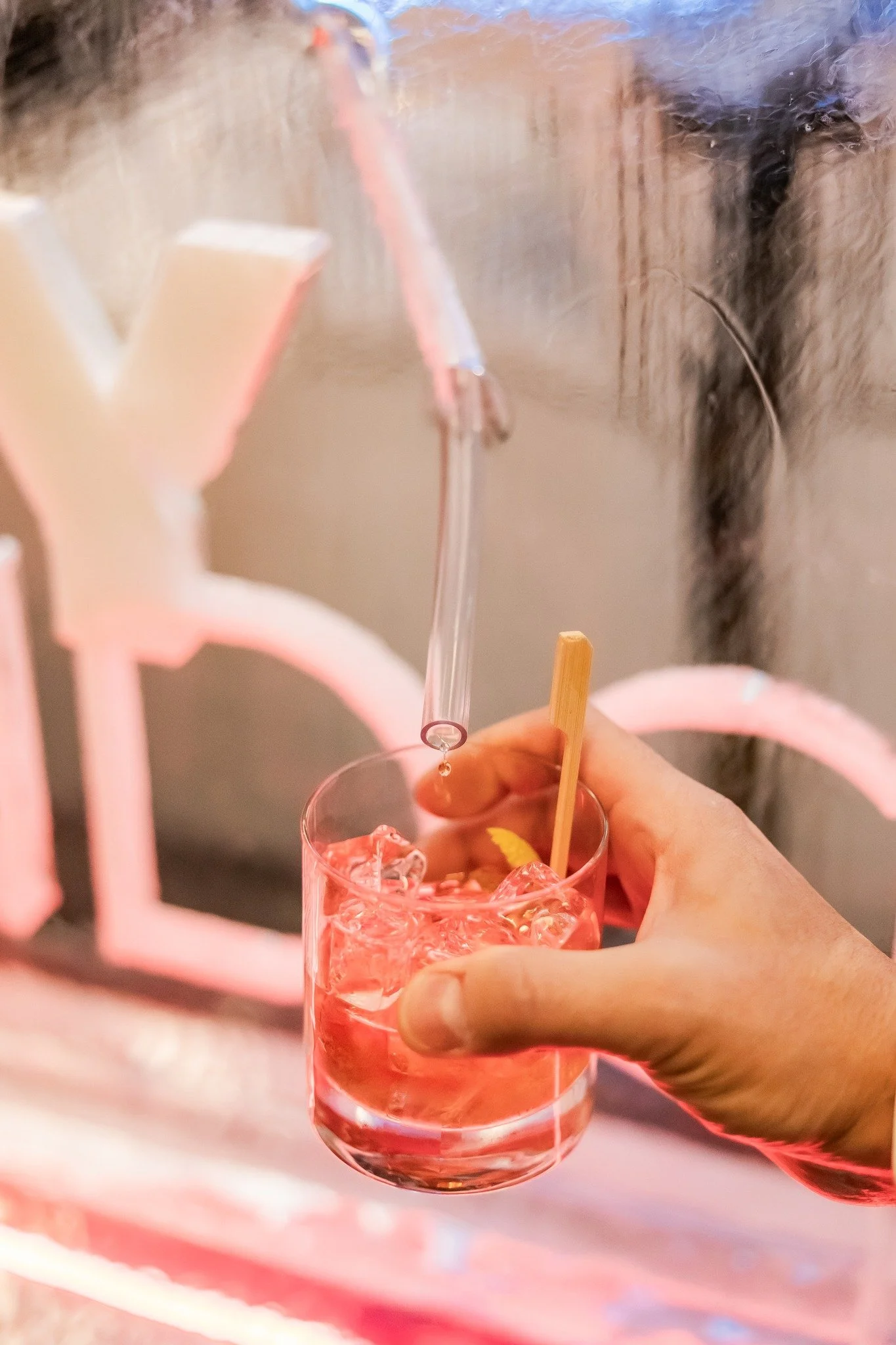 A hand holding a glass of pink drink with ice cubes, a wooden stir stick, and a straw, with a pink neon sign in the background.