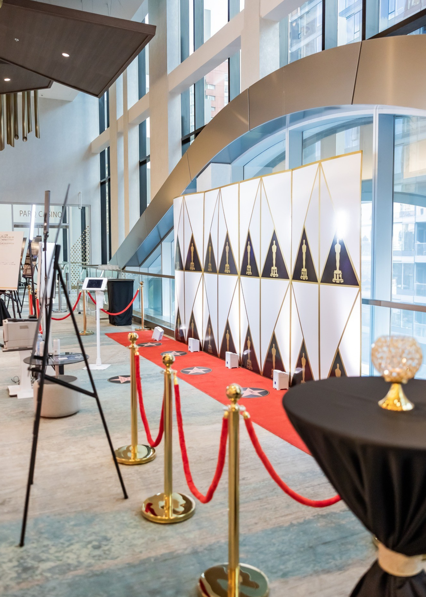 Red carpet with gold stanchions leading to an event backdrop featuring an Oscar statue pattern, inside a modern building with large windows.