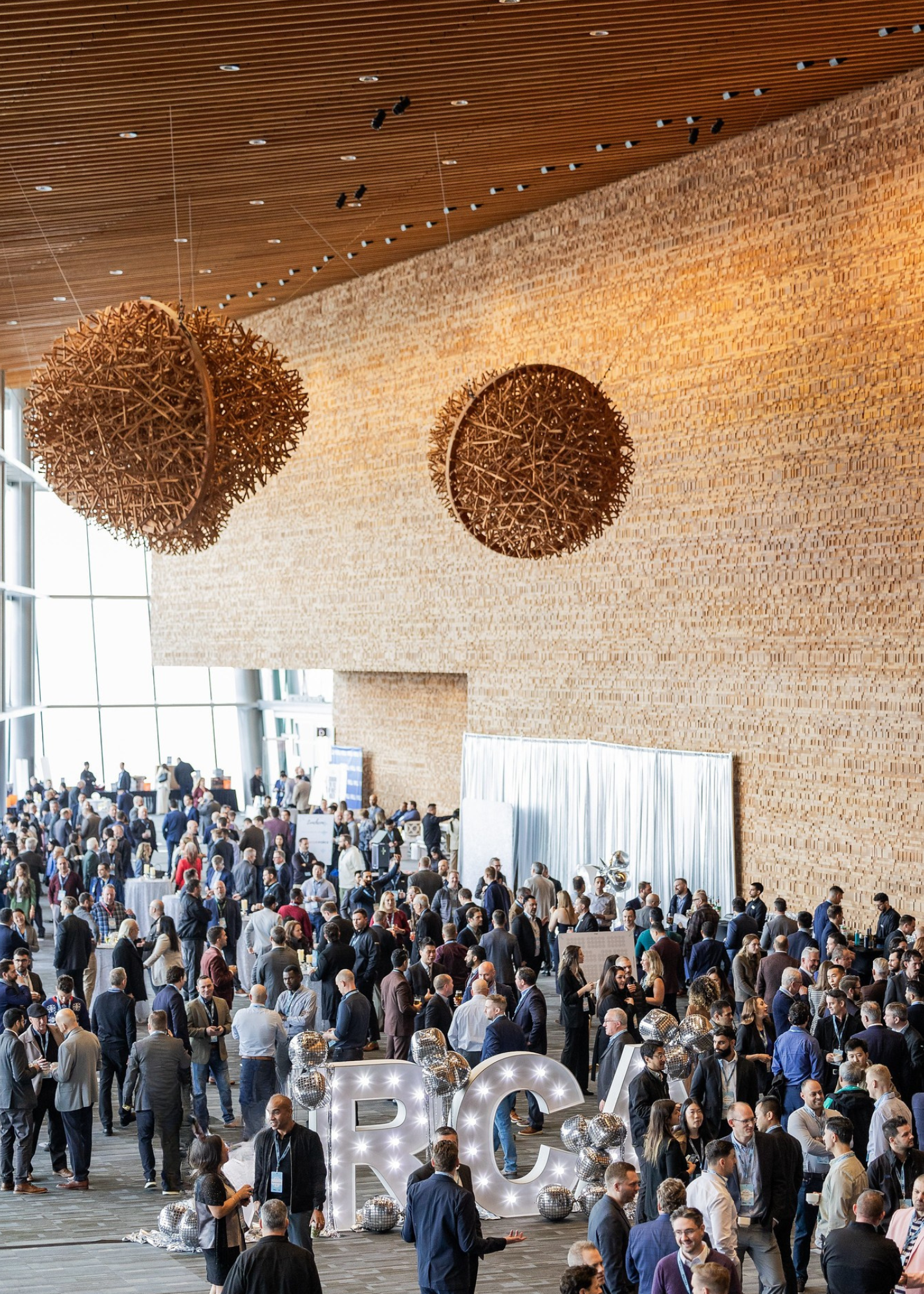 A crowded conference or networking event with many people in business attire in a spacious, modern venue with wooden walls and large pendant lights.