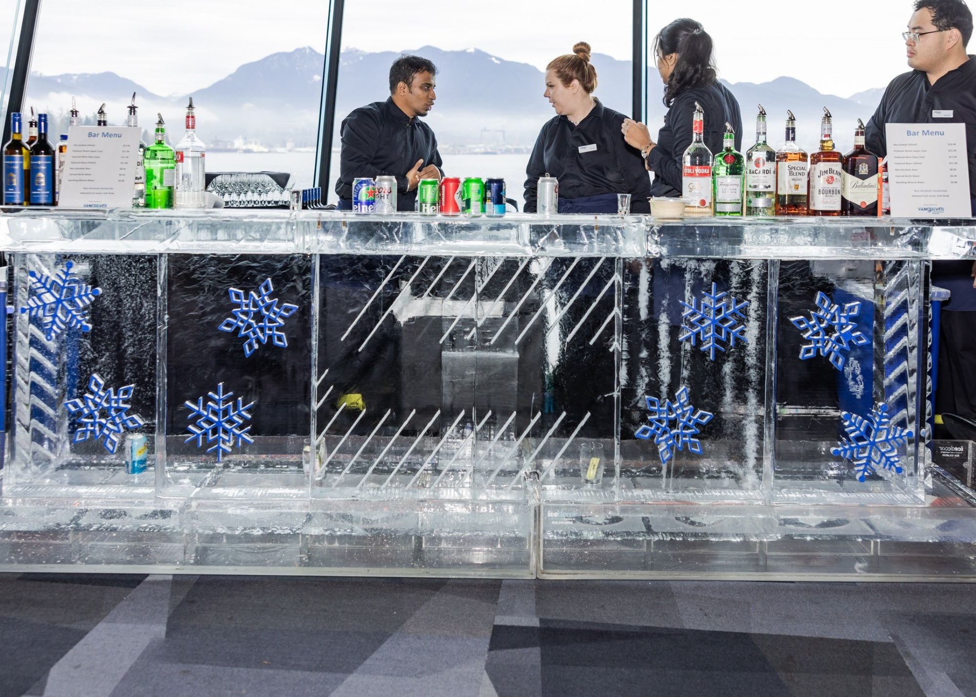 Ice bar with snowflake decorations, bartenders preparing drinks, bottles of liquor and cans of soda on the bar counter, and a scenic mountain view in the background.