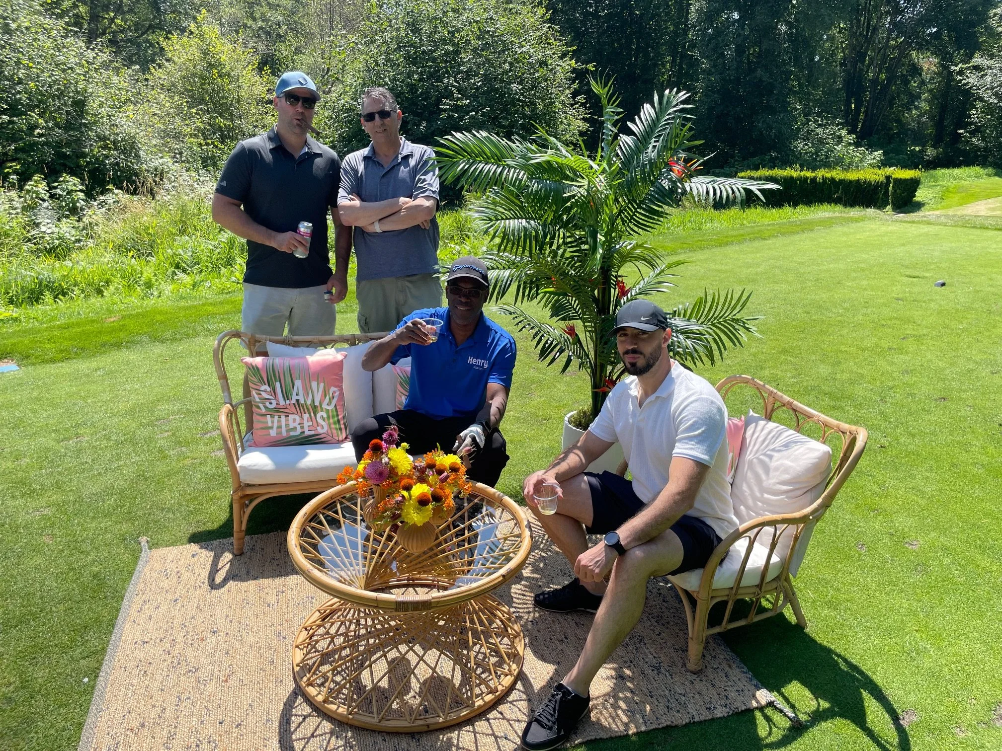 Four men enjoying drinks outdoors in a lush green garden, seated on wicker furniture with tropical-themed pillows and a flower arrangement, with tall trees and a well-maintained lawn in the background.