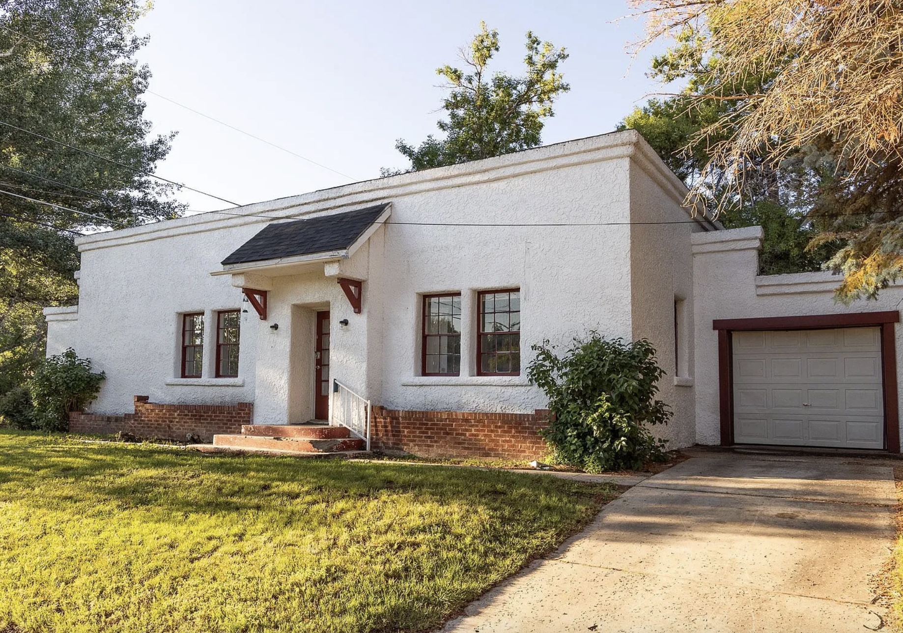 A small, white stucco house with red trim, brick accents, and a black roof. The house has three windows, a front door with steps, and a driveway leading to a garage. There is a green lawn and trees around the house.