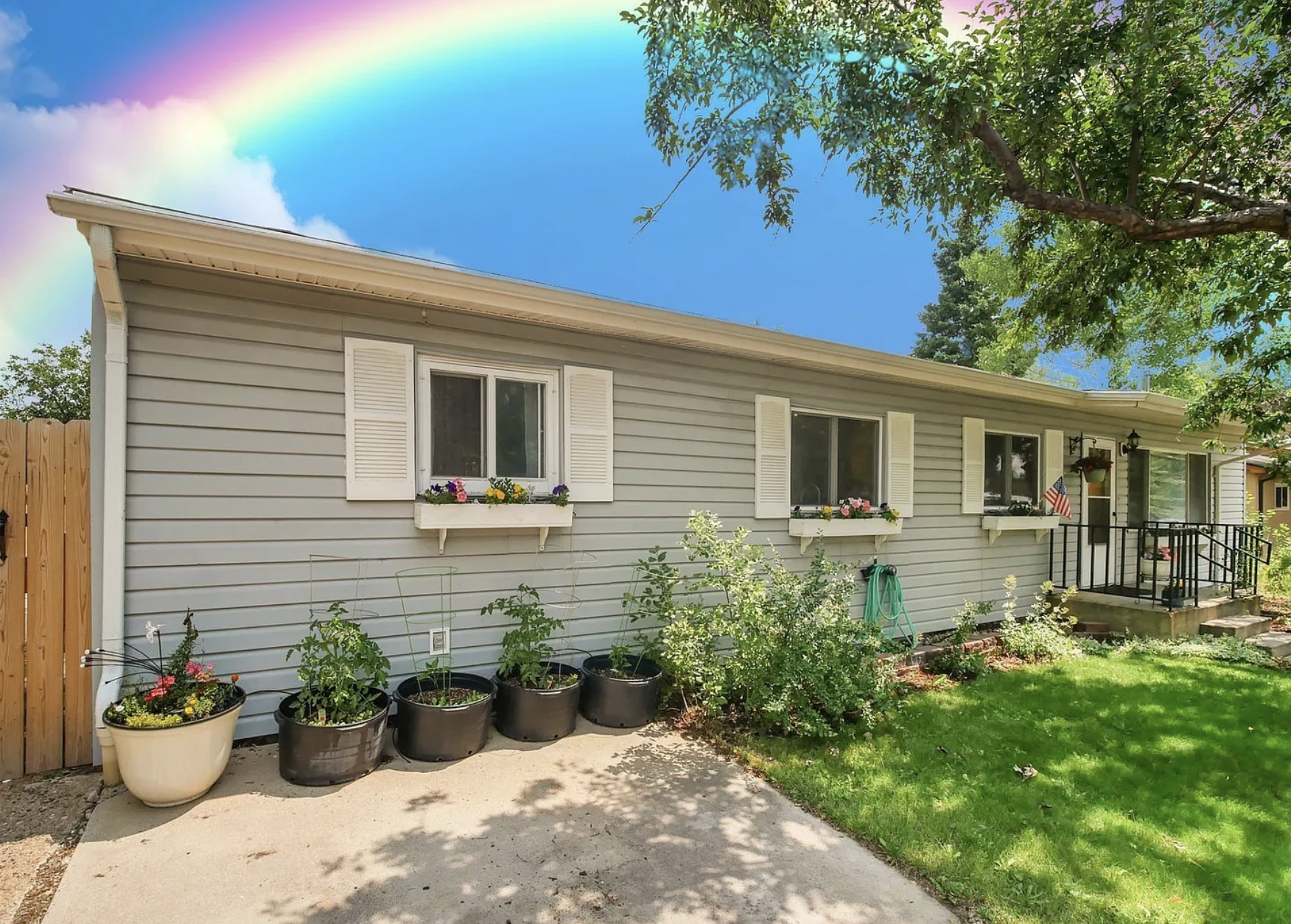 A single-story house exterior with beige siding, white shutters, flower boxes under windows, a small porch with American flag, potted plants, a garden hose, and a vibrant rainbow in the sky overhead.