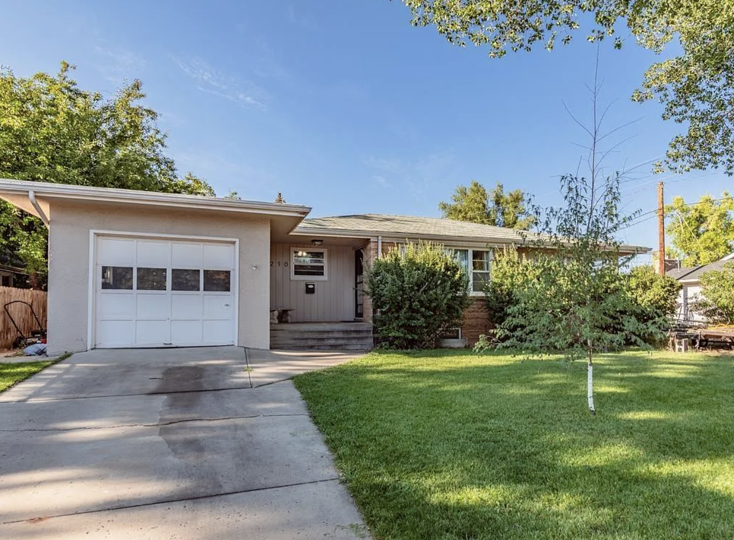 Front view of a single-story house with a garage, concrete driveway, and a well-manicured lawn with small trees and bushes, under a clear blue sky.