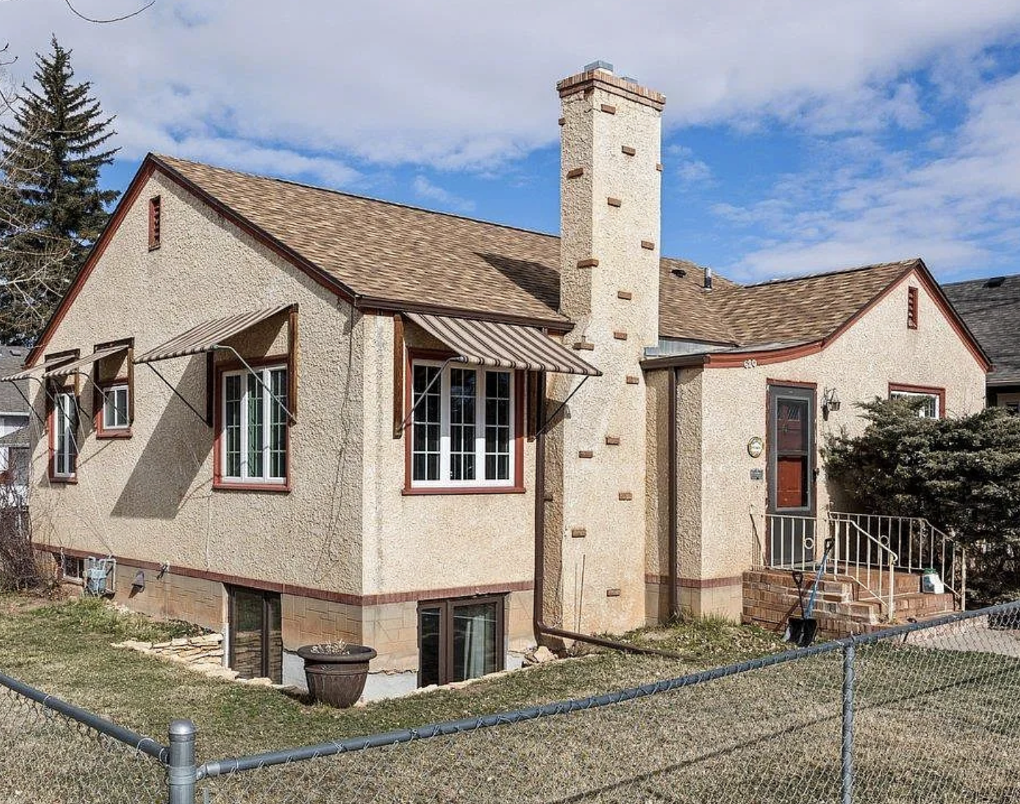 A two-story house with beige stucco exterior, brown roof shingles, and a chimney. It has striped window awnings over the front windows, a small set of stairs leading to the front door, and a fenced yard.