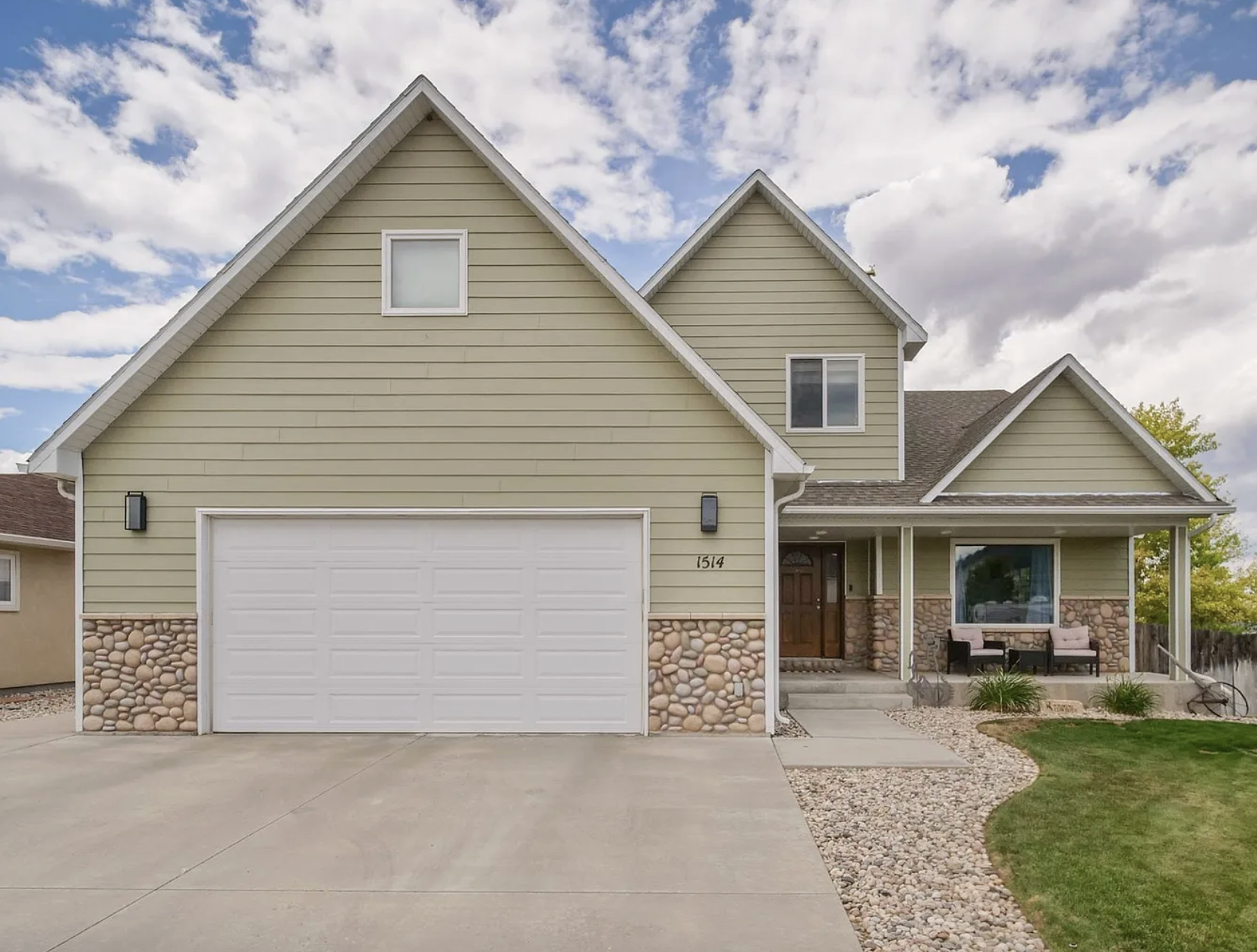 Front view of a two-story suburban house with a light green exterior, stone accents, a white garage door, and a covered porch with seating. The house has multiple gable roofs and a well-maintained lawn with a gravel pathway.