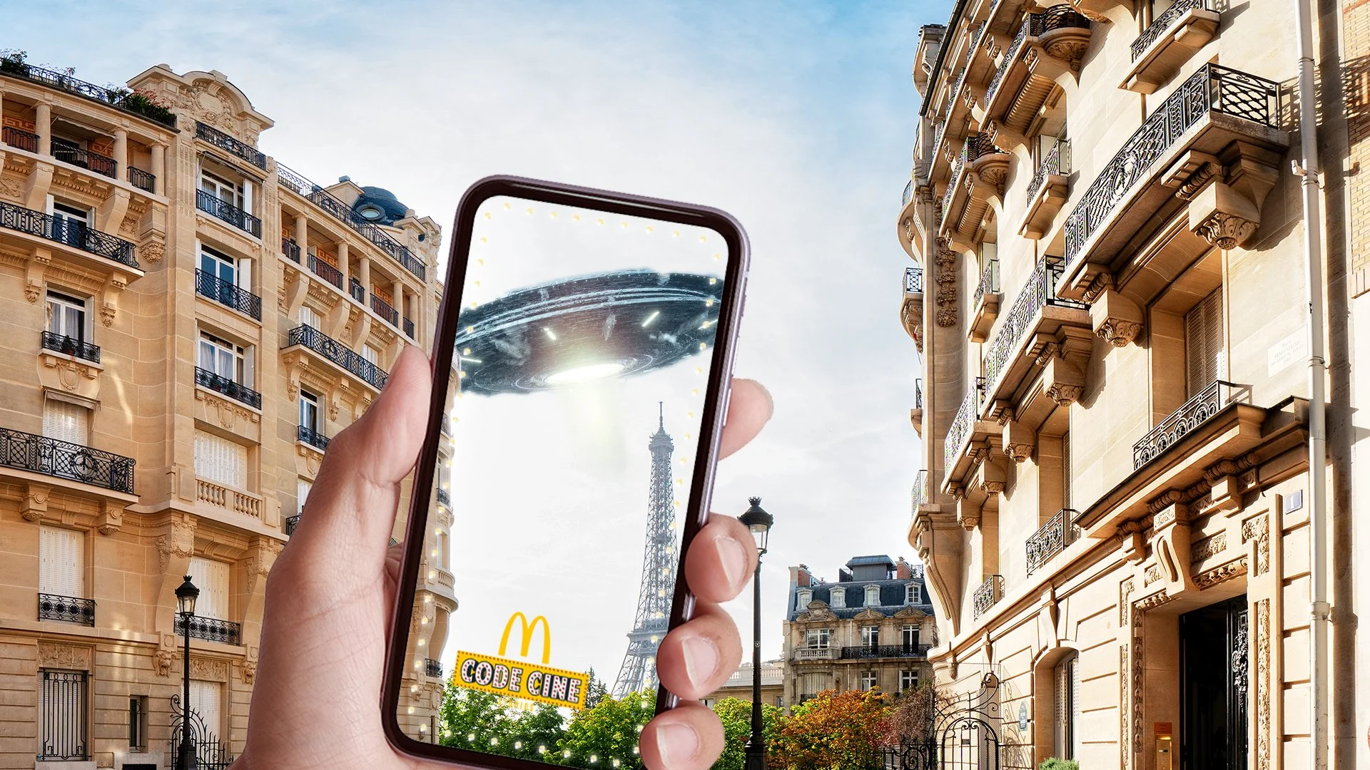 Un téléphone portable dans une main en train de prendre une photo de la Tour Eiffel avec un OVNI dans le ciel, dans une rue parisienne ensoleillée.