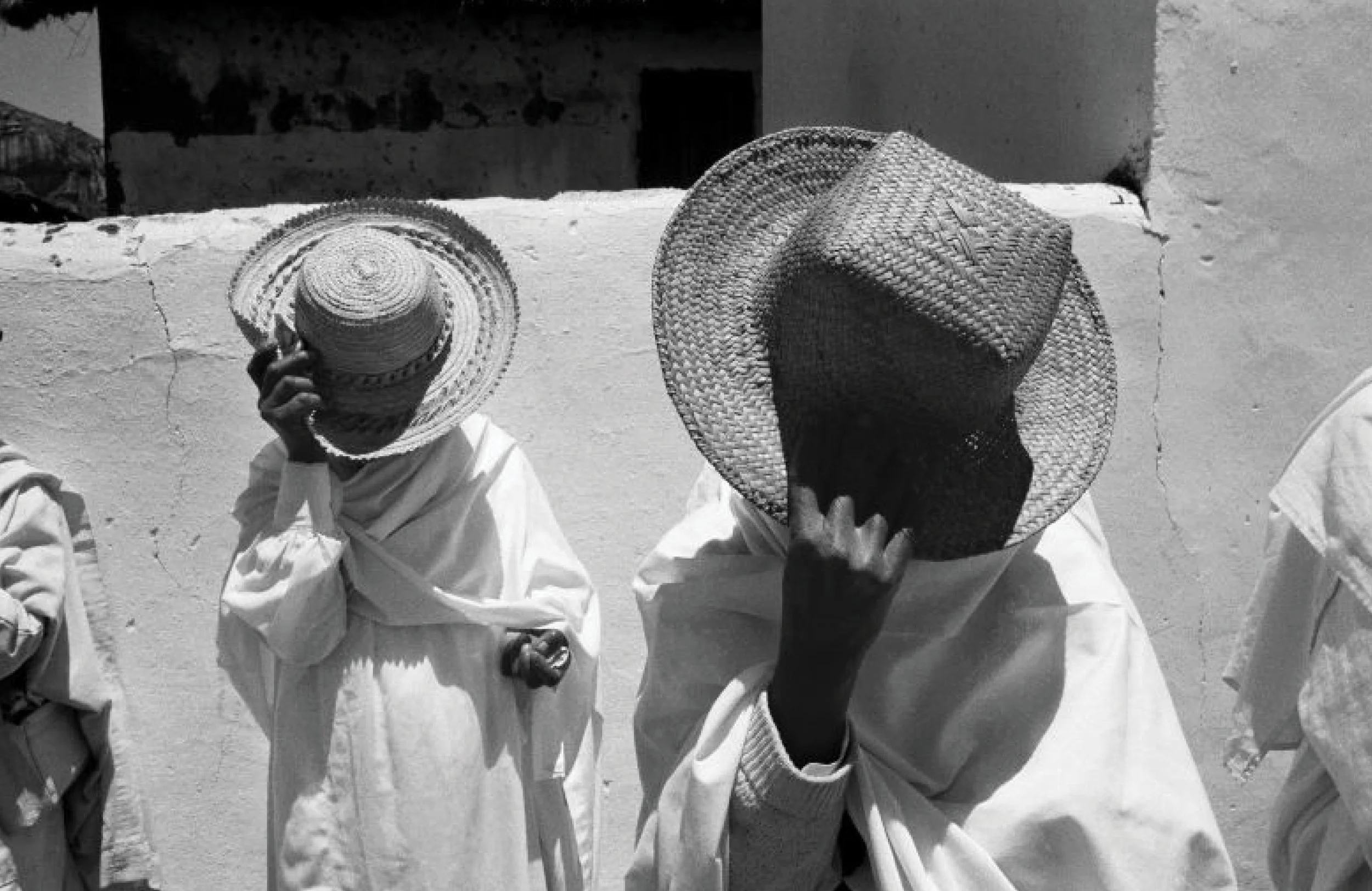 Deux personnes portant des vêtements traditionnels et des chapeaux tressés, debout côte à côte, dans un environnement extérieur en pierre ou en adobe.