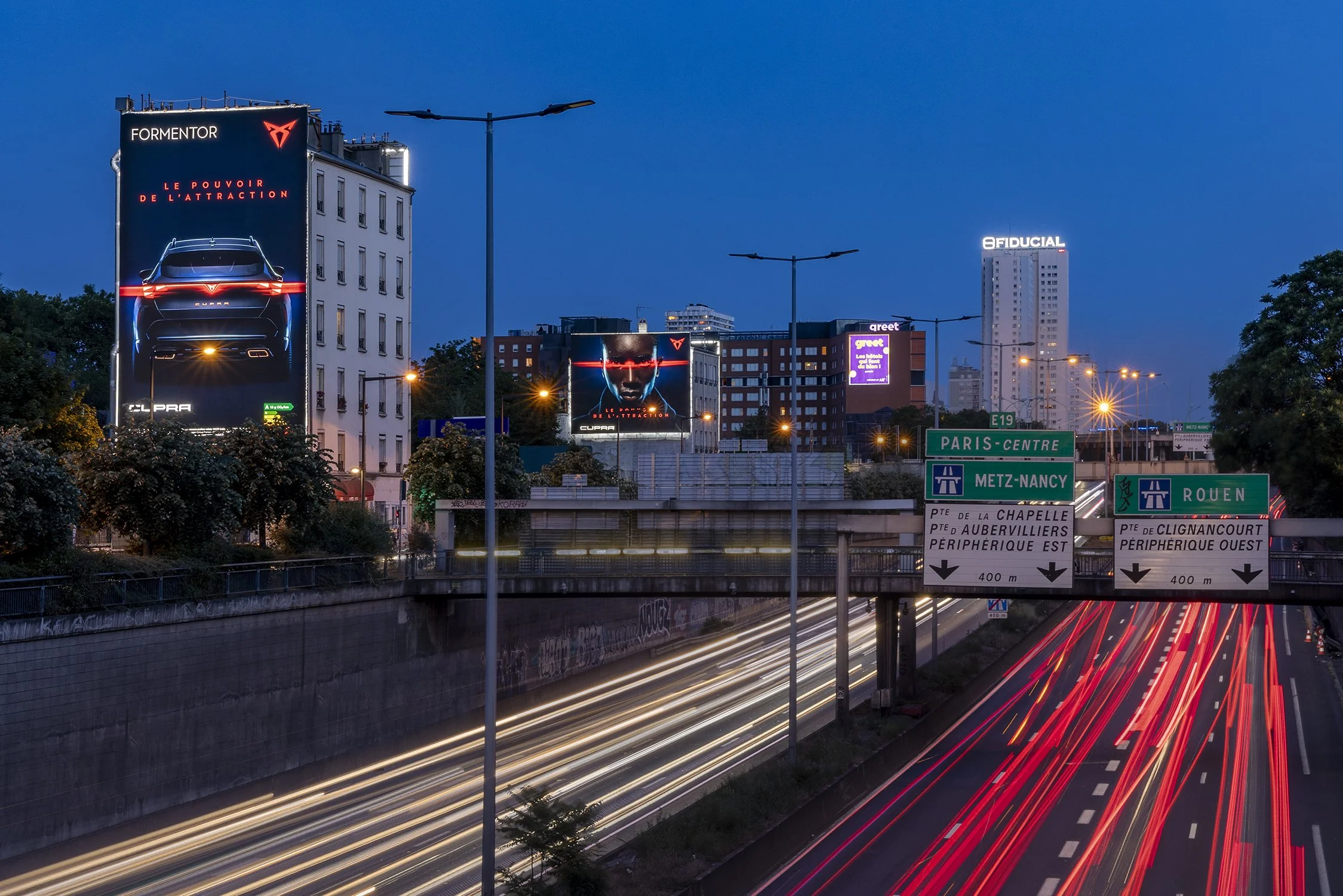 Une photo urbaine prise en soirée montrant des panneaux publicitaires illuminés, des bâtiments et des routes à trafic long exposure avec des trainées lumineuses de véhicules, ainsi que des panneaux de signalisation routière indiquant différentes directions