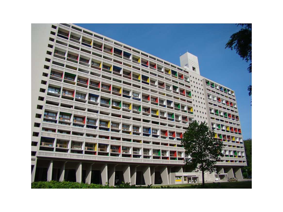 Immeuble moderne avec façades colorées et balcons, sous un ciel bleu, avec un arbre devant.