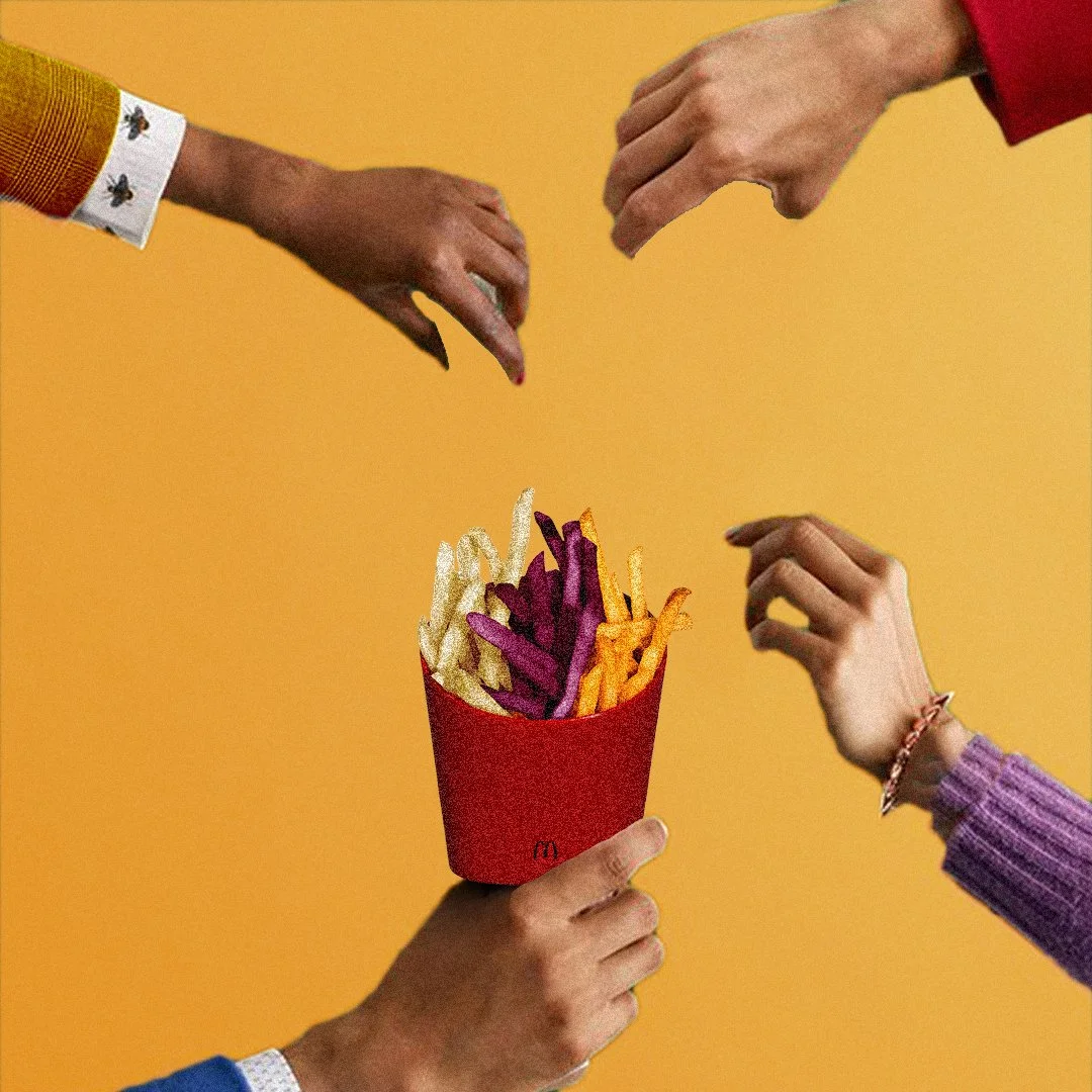 Une main tient une grande boîte de frites colorées (blanches, violettes, oranges) avec un fond orange vif, entourée de mains de différentes personnes.