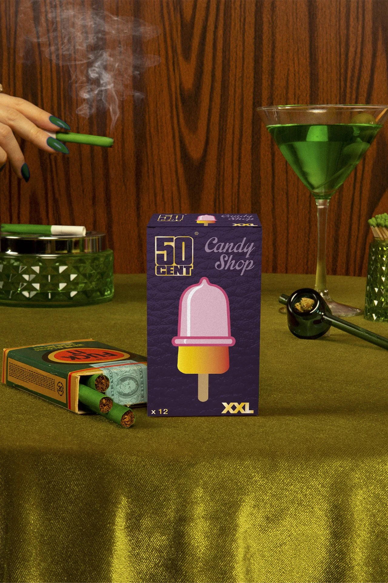 Une table avec une boîte de feux d'artifice de 50 centimes, une boîte de cigarettes, un verre à martini avec un cocktail vert, une cigarette allumée, un pipe à tabac, et des cigares.