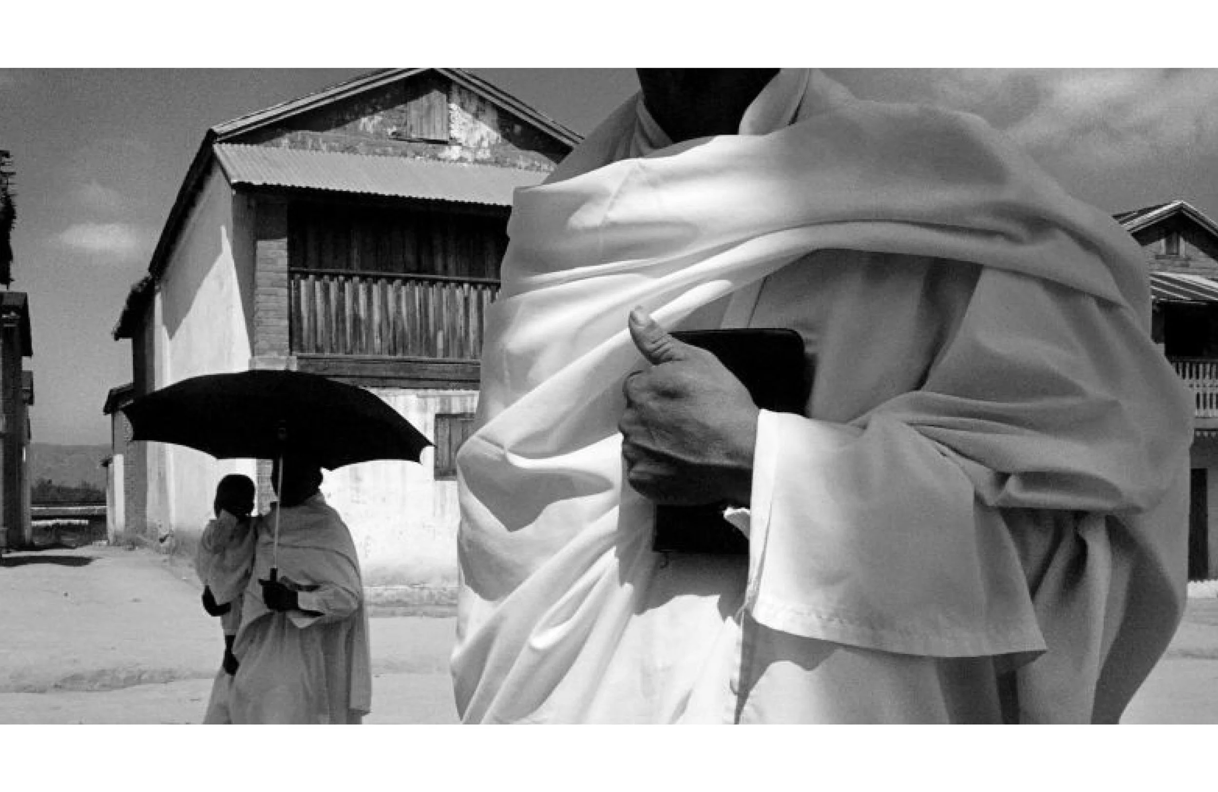 Une personne portant une robe ou un vêtement ample blanche, tenant une pièce ou un objet dans sa main, avec deux autres personnes en arrière-plan, dont une tenant un parapluie, dans une scène extérieure.