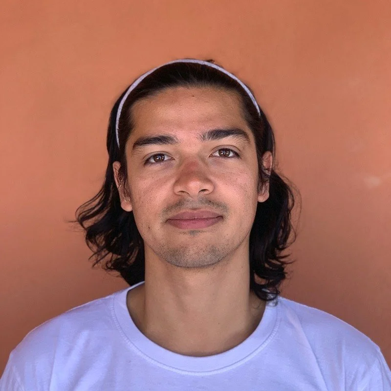 Un jeune homme avec des cheveux longs, portant un bandeau blanc et un t-shirt blanc, se tient devant un mur orange.
