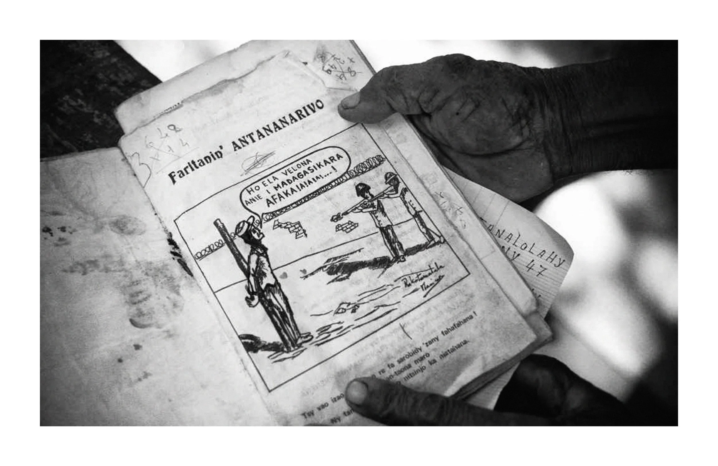 Une main d'homme tient une vieille bible ouverte. La page montre un dessin illustrant deux personnes avec des bâtons et un texte en anglais. La photo est en noir et blanc.