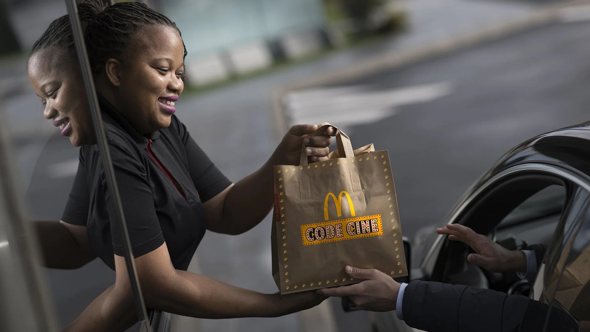 Une employée de McDonald's donne un sac contenant un film, avec le logo McDonald's et la mention "Code Cine", à un client dans une voiture.