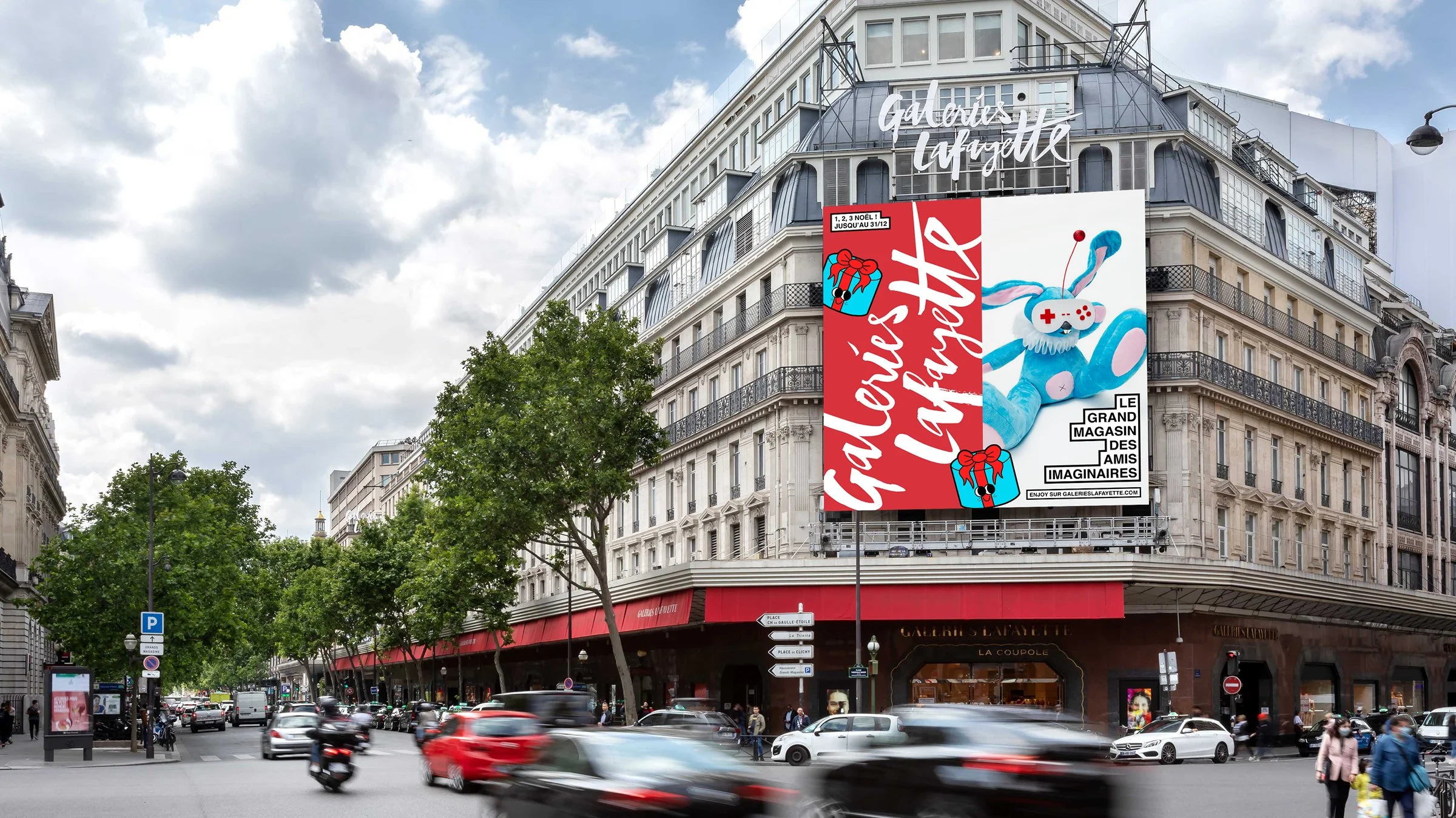 Grand bâtiment parisien avec une grande affiche publicitaire pour Galeries Lafayette, montrant un pouf bleu en forme de lapin avec une manette de jeu vidéo et un hérisson en peluche. Le ciel est partiellement nuageux, et beaucoup de voitures et de piétons sont visibles.