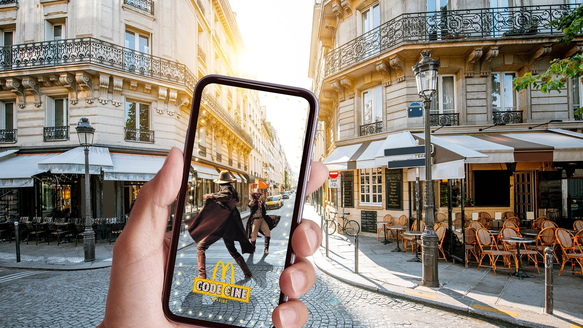 Une personne tient un téléphone qui affiche une scène de rue parisienne avec des passants et des cafés, tandis que le paysage réel montre la même rue. La scène capture une intersection avec des bâtiments en style haussmannien, des lampadaires et des 