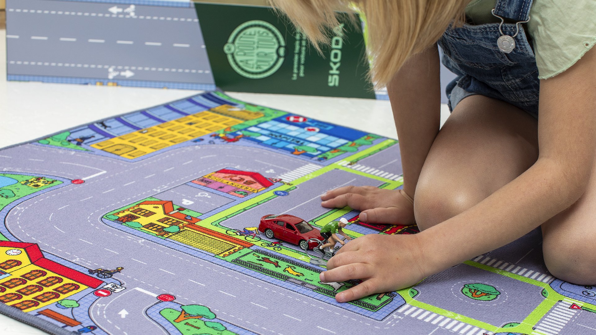 Enfant jouant à un jeu de société avec un tapis illustrant une ville, avec des voitures miniatures, une figurine de cycliste et d'autres éléments de jeu sur le tapis.
