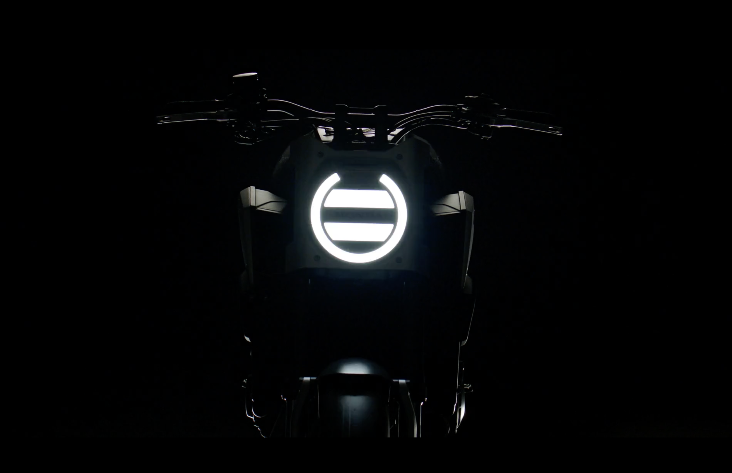 Vue frontale d'une moto électrique en mode sombre, avec une signature lumineuse blanche en forme de cercle sur le devant et des lignes lumineuses sur le guidon.