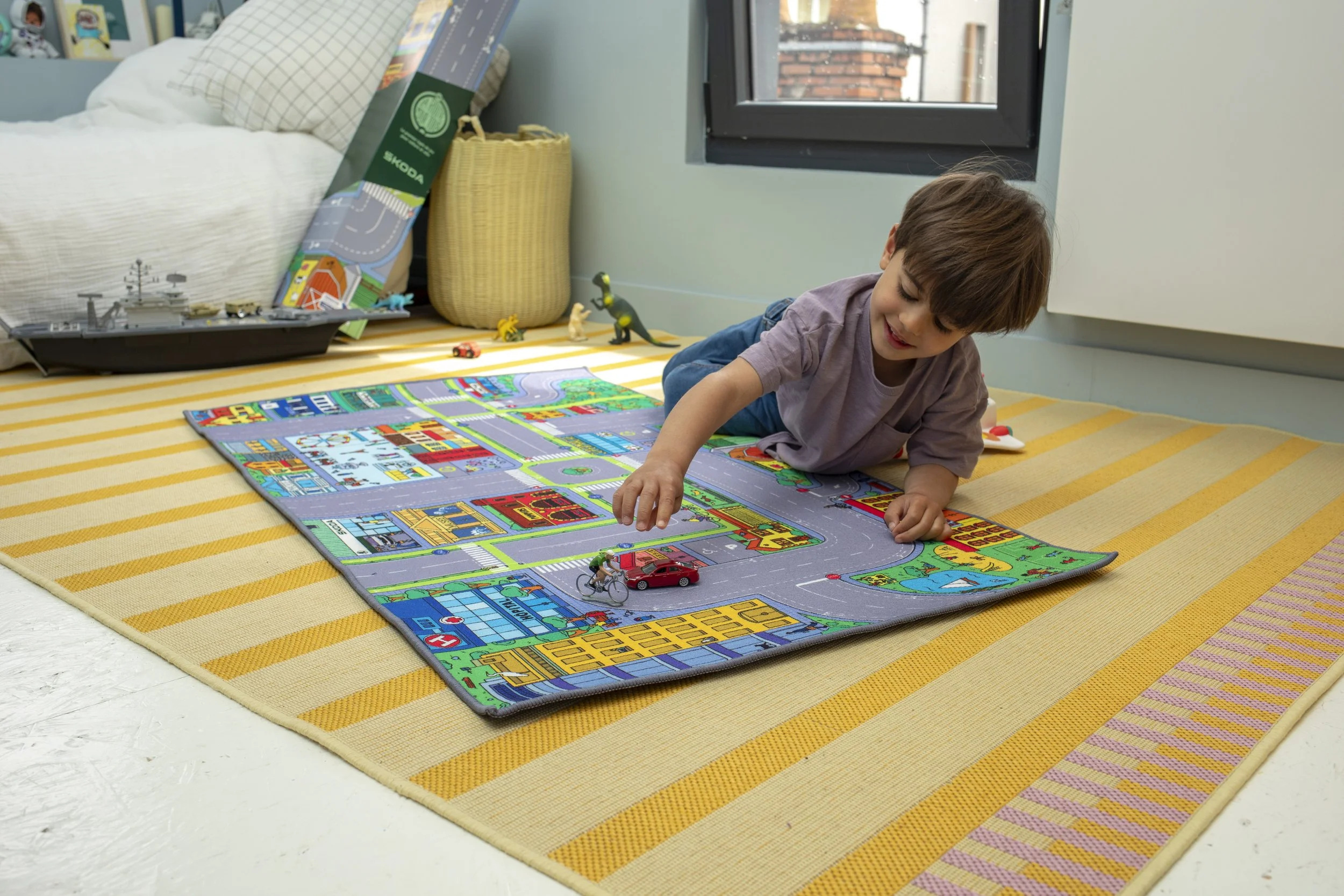 Un garçon joue avec un tapis de jeu de ville avec des voitures Miniatures dans une chambre. Il est allongé sur un tapis jaune rayé, près d'une fenêtre, avec une cabane en osier et des jouets à l'arrière.