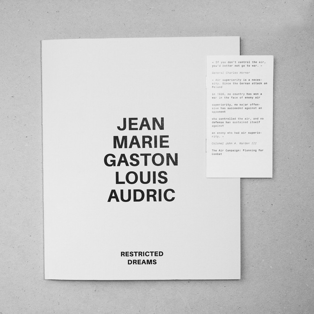Couverture d'un livre intitulé "Restricted Dreams" avec le nom de l'auteur "Jean Marie Gaston Louis Audric" en gros caractères noirs sur fond blanc, posé sur une surface grise, avec une petite feuille de papier à droite contenant des citations et des