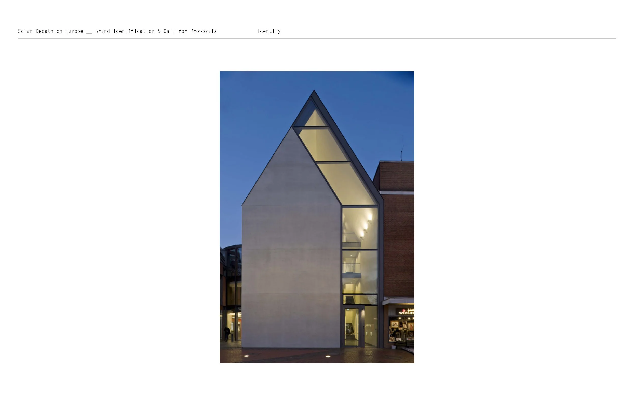 Une façade de bâtiment moderne avec de grandes fenêtres et un toit en pente, éclairée par des lumières dans une zone urbaine, en soirée.