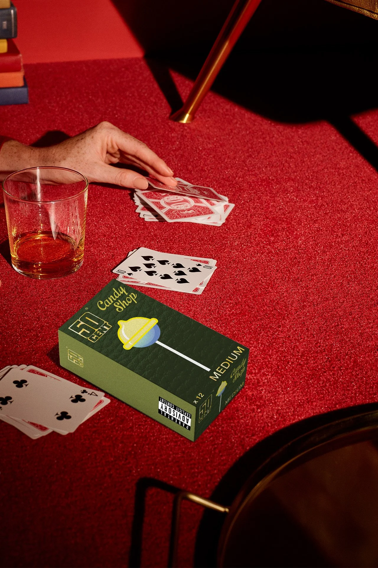 Une table rouge avec un verre de boisson, un paquet de jeux de cartes, et quelques cartes posées sur la table. Un bras est en train de jouer avec les cartes.