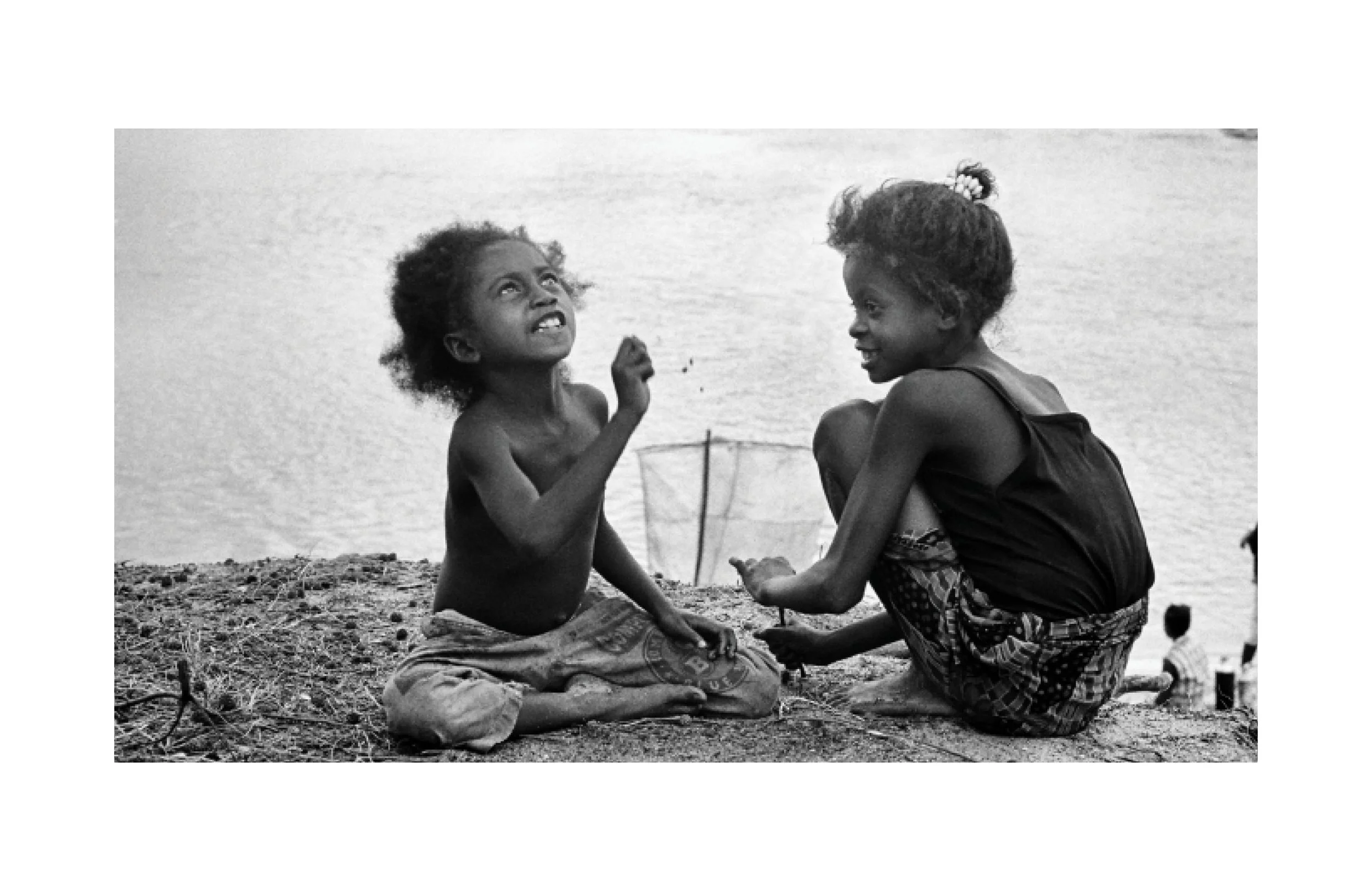 Deux jeunes enfants, assis sur la plage, parlent. L'un regarde vers le haut et l'autre le regarde en souriant.