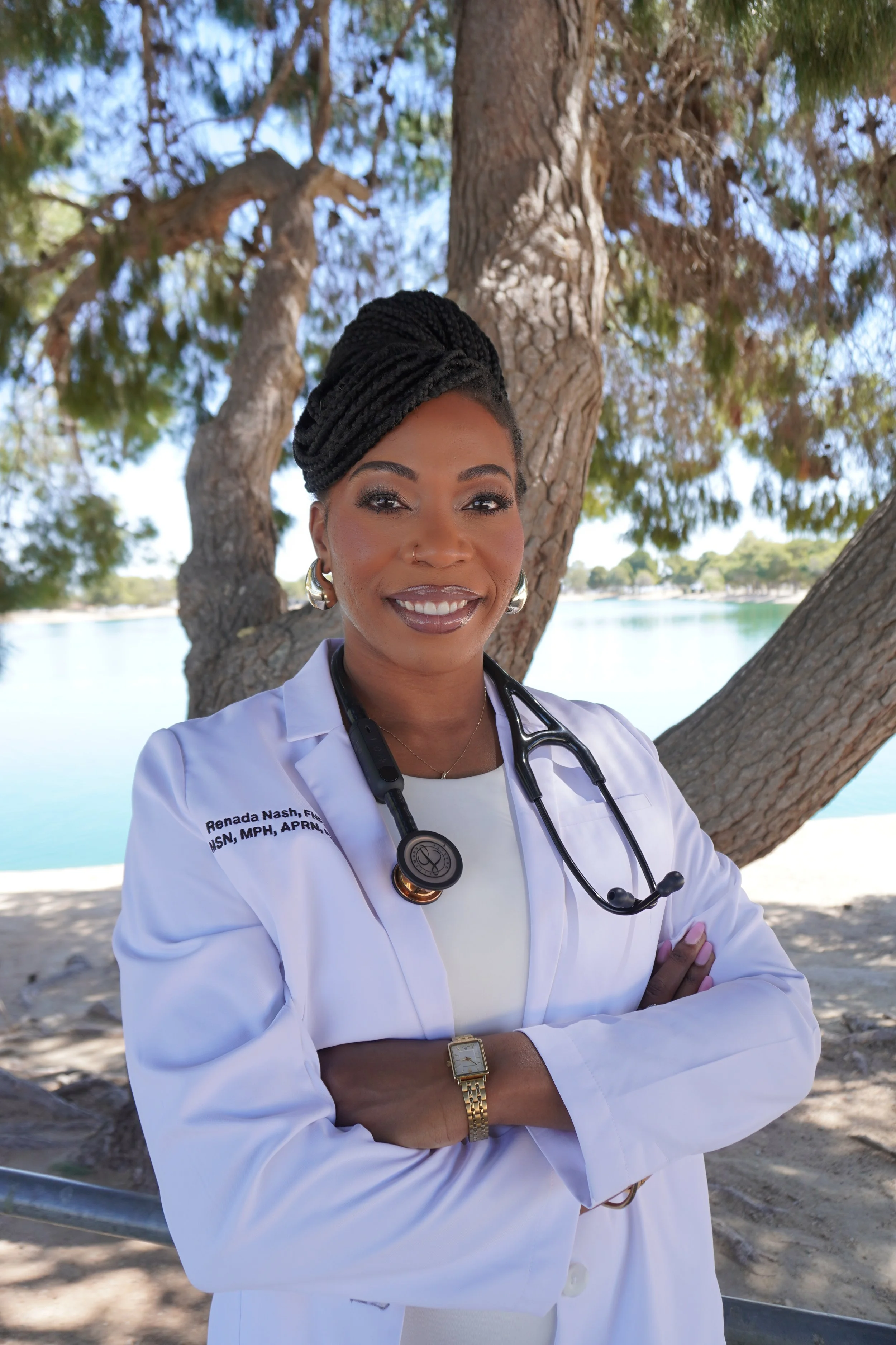 A female doctor standing outdoors near a large tree, with water and a distant shoreline in the background. She has braided hair, wears a white medical coat with her name and credentials, a stethoscope around her neck, earrings, a wristwatch, and a bracelet, and is smiling with her arms crossed.