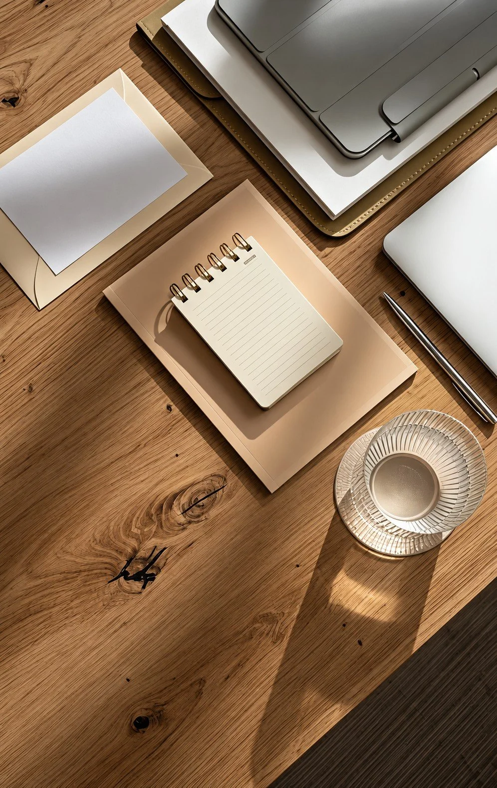 Overhead view of an organized workspace with a notebook, laptop, pen, and glass of water on a sunlit wooden desk.