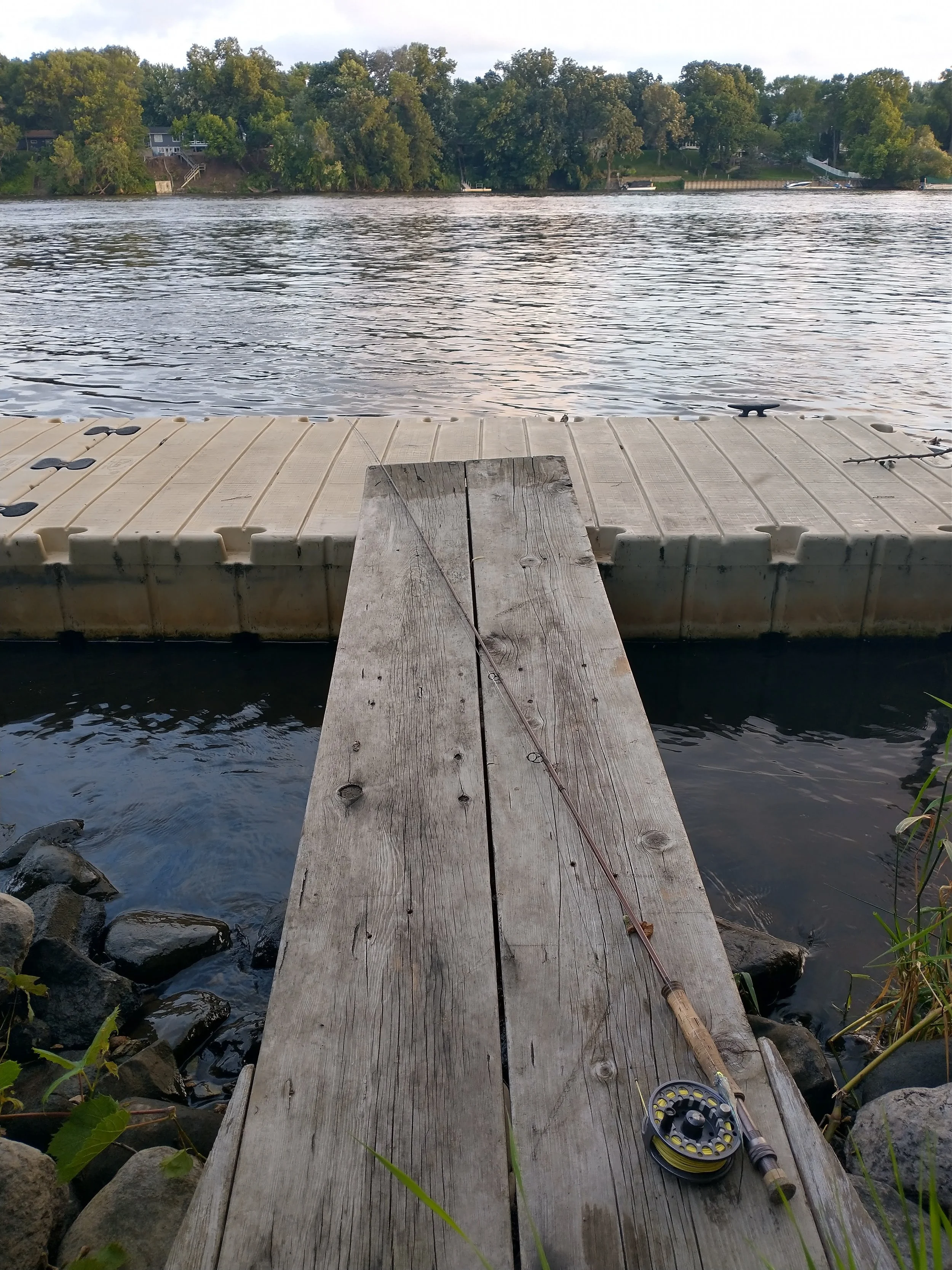 A wooden dock extends into a lake with a fishing rod resting on the dock. The lake is calm, with trees and houses visible on the far shore.