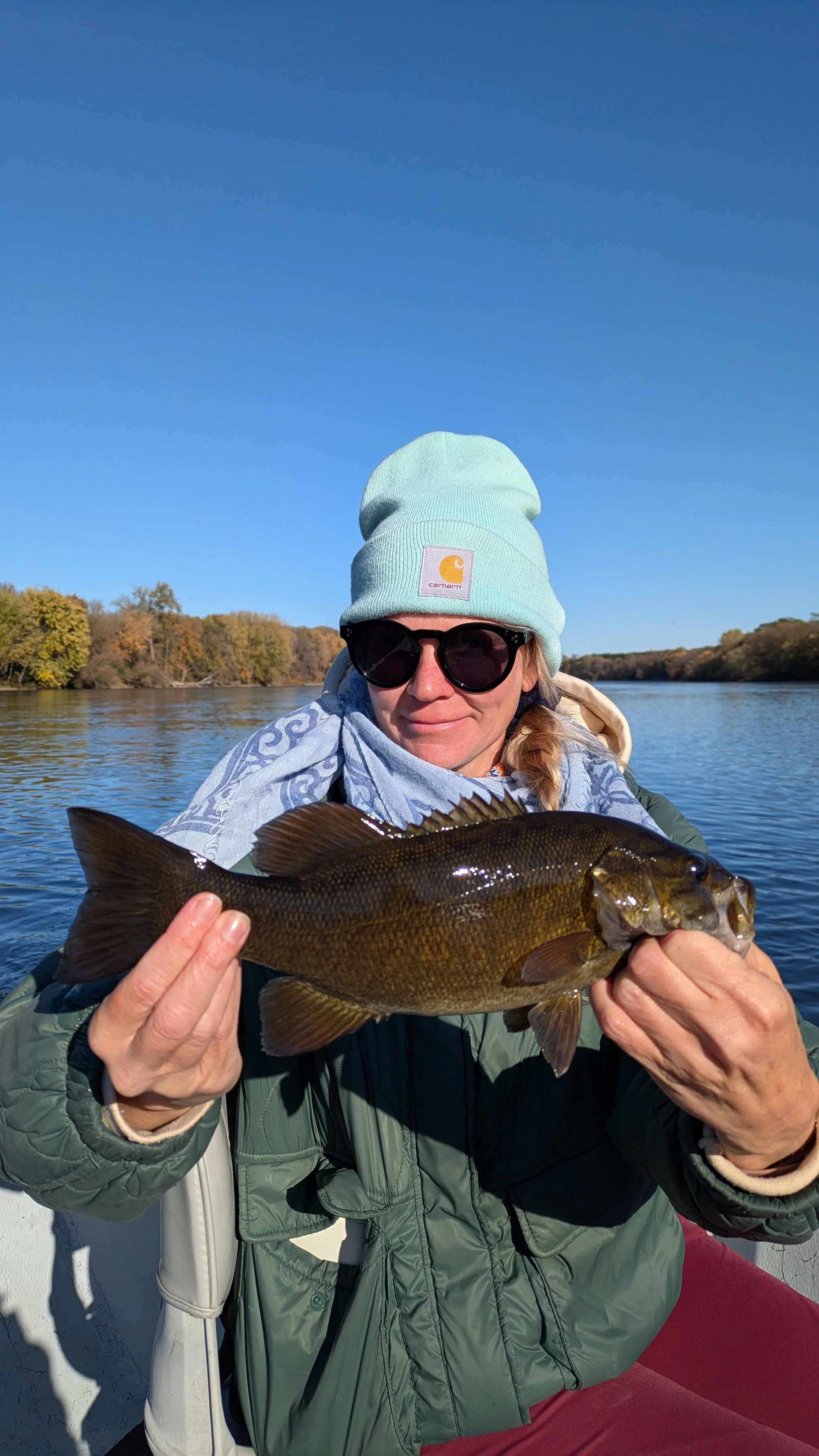 A woman wearing a light blue beanie, sunglasses, and a green jacket holding a fish on a boat in a river with trees in the background.