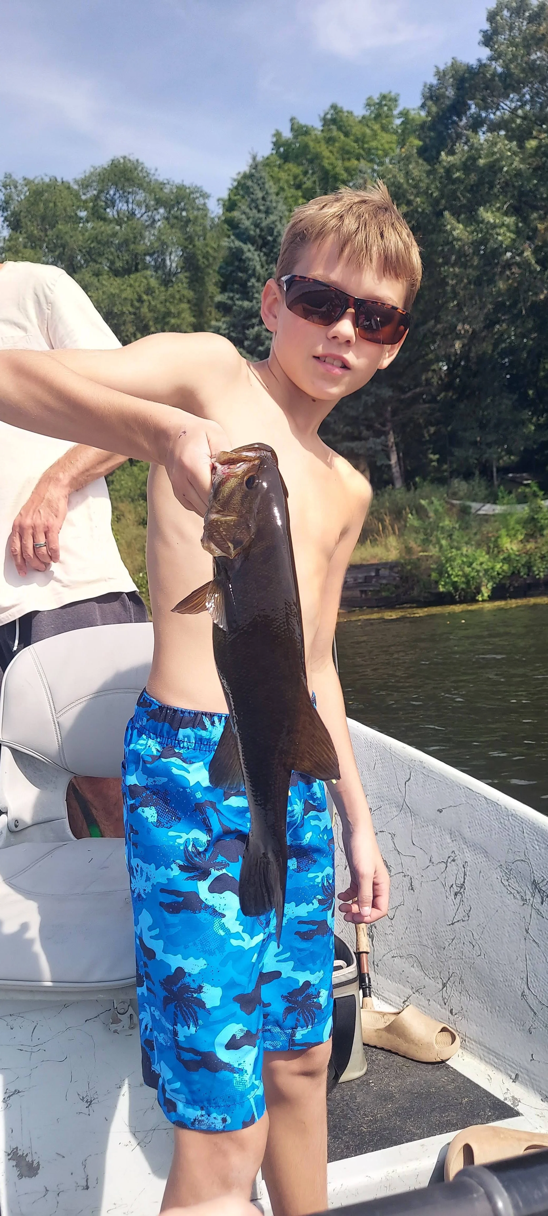 A young boy wearing sunglasses and blue camouflage swim trunks holding a large fish on a boat during a sunny day.