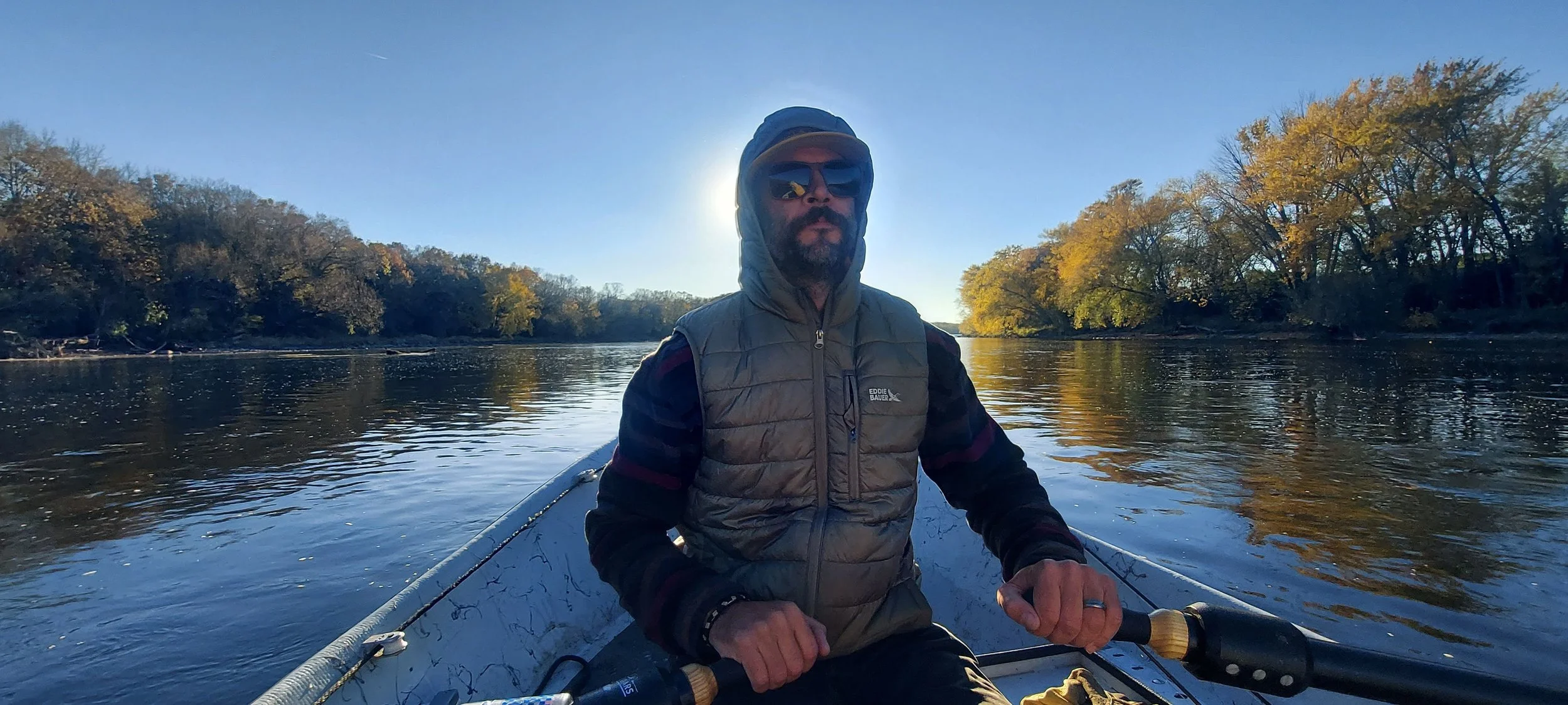 A man wearing sunglasses, a hat, and a puffy vest is sitting in a boat on a river with trees showing fall foliage on both sides, under a clear blue sky during daytime.